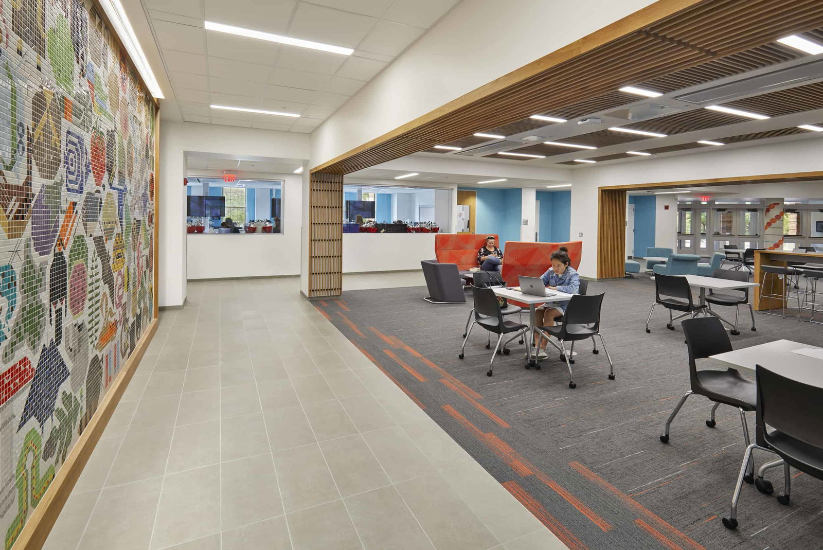 This modern study and lounge area is a dynamic hub for student collaboration and quiet focus. Featuring a striking mosaic wall of colorful, abstract shapes and symbols near the entrance, the space immediately conveys a creative, energetic vibe. Open seating areas include a mix of high-backed, semi-private booths in bright red tones, soft lounge chairs, and moveable tables with ergonomic chairs — perfect for group work or solo study. Wood-slat ceiling details and carefully chosen accent colors like sky blue and deep orange give the space warmth and character, while natural light flows in from large windows. The overall design balances form and function, fostering both academic productivity and casual interaction.