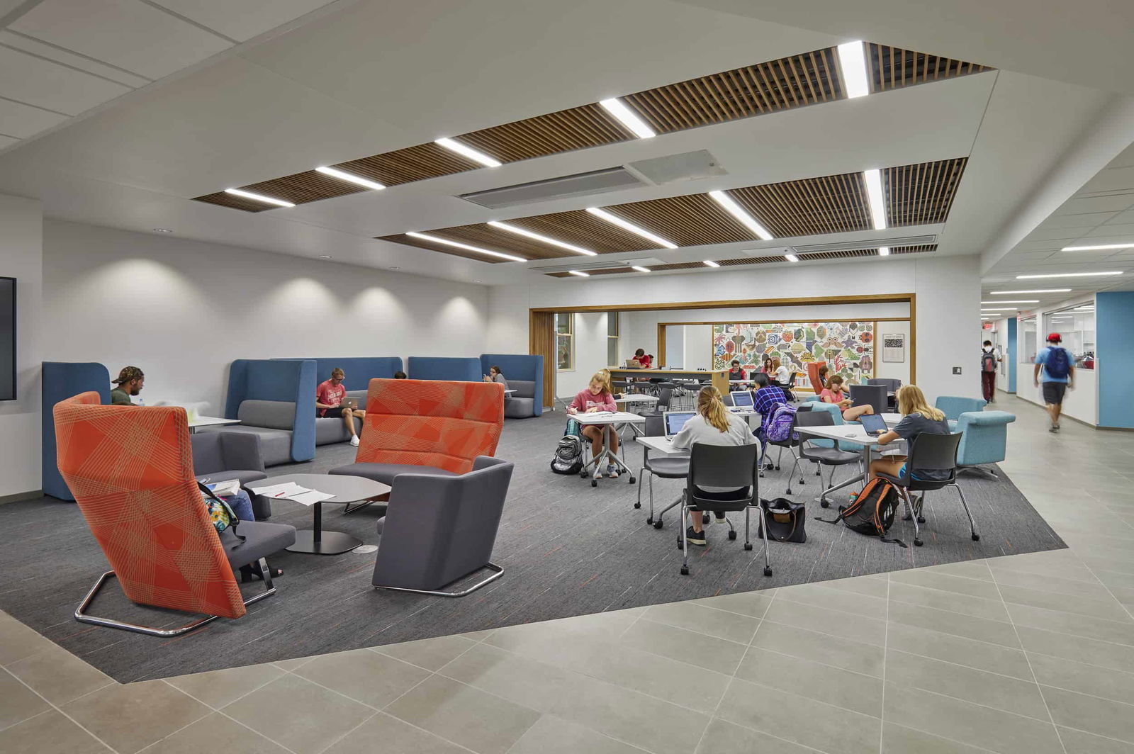 Designed to foster academic collaboration and flexible learning, this open-concept student commons is a vibrant extension of the building’s educational mission. The area features a mix of seating arrangements—from individual study tables to cozy lounge nooks with high-back chairs in bold orange and deep blue. The ceiling integrates stylish wood paneling, creating both visual interest and acoustic comfort. Students can be seen actively engaged with laptops, group discussions, or independent work, demonstrating the space’s adaptability. A colorful mosaic wall provides a creative focal point, reinforcing the building’s emphasis on science, diversity, and innovation.
