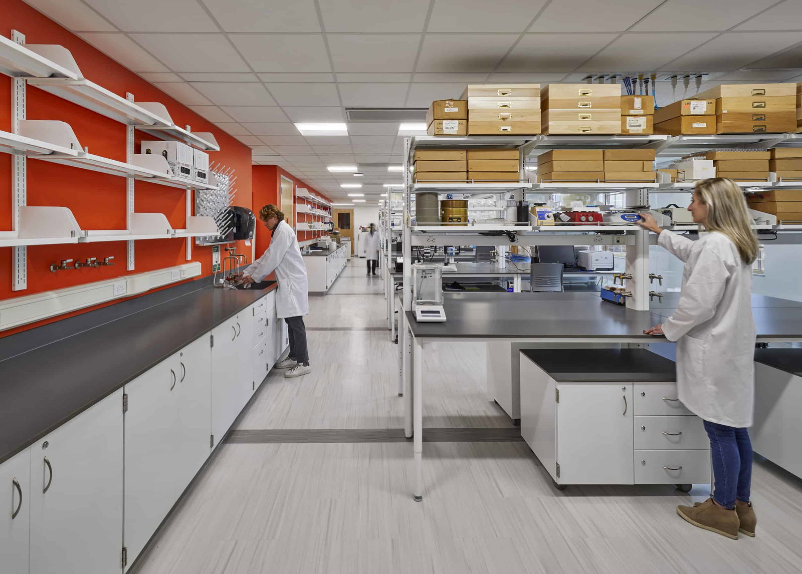 This image shows a modern scientific laboratory space. Two researchers in white lab coats are working at different stations - one person on the left side at a counter with an orange accent wall behind it, and another person on the right side at a workstation with laboratory equipment. The lab features extensive white cabinetry with dark countertops, metal shelving systems holding numerous storage boxes and scientific instruments, and light-colored wood-look flooring. The space is well-organized with various scientific equipment visible on the counters and shelves. The bright orange wall adds a distinctive pop of color to the otherwise clean, clinical environment. In the background, another person in a lab coat can be seen walking through the space. The laboratory appears to be designed for scientific research with ample storage and work surfaces.