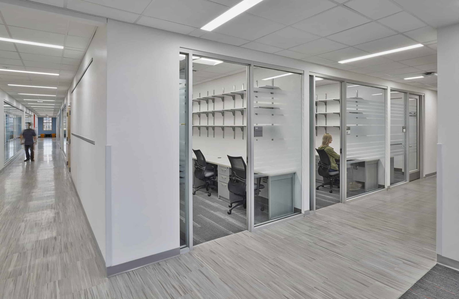 This image shows a modern academic or office space with glass-walled rooms. In the foreground is a small office or study room with glass walls and doors, containing a white desk with two black office chairs and multiple wall-mounted shelves. A person can be seen sitting at one of the workstations inside this glass-enclosed space. The surrounding area features light gray wood-look flooring and white walls with recessed linear lighting in the ceiling. To the left, there's a corridor where another person can be seen walking in the distance. The overall design has a clean, minimalist aesthetic with ample natural light and visibility between spaces, typical of contemporary educational or research buildings.