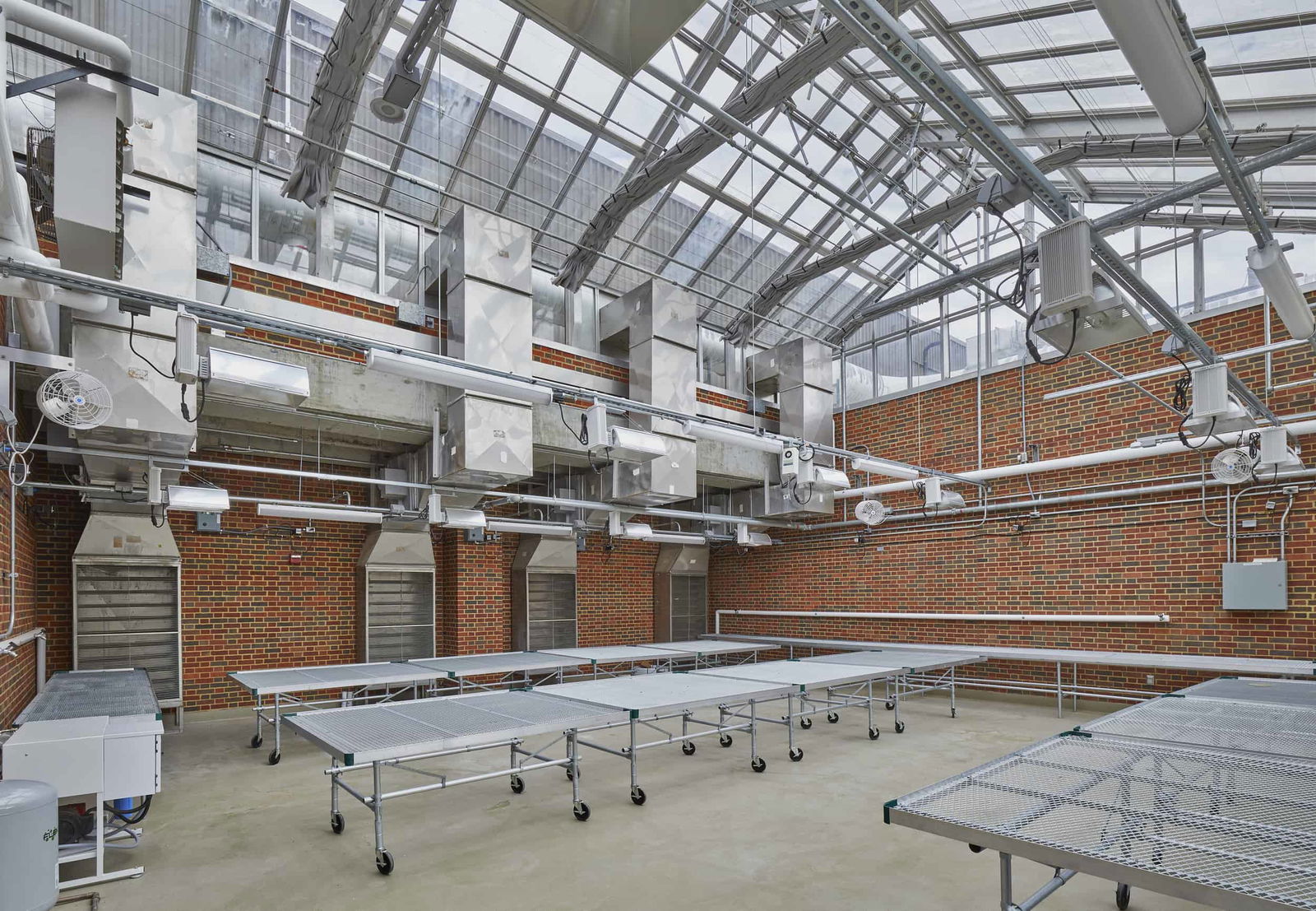 This image shows a specialized work area in the Hughes and Pearson Hall at Miami University. The space features brick walls and an extensive glass ceiling with a metal framework structure that allows natural light to enter. The room is equipped with multiple long, metal mesh tables on wheels that appear to be designed for research purposes. Above the tables, there's sophisticated overhead equipment mounted to the ceiling, including what look like ventilation systems, lighting fixtures, and possibly automated watering or climate control mechanisms. The space has a concrete floor and appears to be designed for scientific research or agricultural studies.