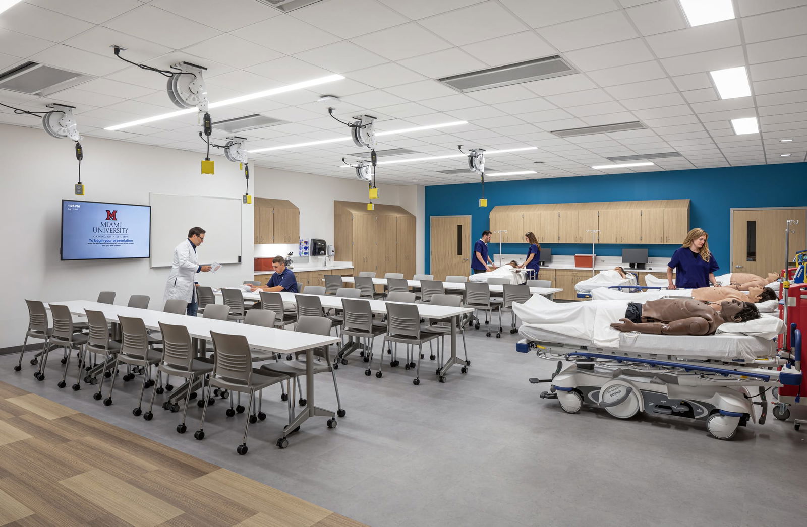 A multipurpose clinical education room in Miami University's Health Sciences Building combining classroom and simulation spaces. On the left, rows of white tables with gray chairs face a monitor displaying the Miami University logo, where an instructor in a white coat reviews materials with a seated student. The right side features hospital beds with medical training mannequins where students in blue scrubs practice patient care techniques. The room includes ceiling-mounted utility reels for medical equipment, wooden storage cabinets against a blue accent wall, and specialized teaching equipment. This integrated learning environment allows for seamless transition between didactic instruction and hands-on clinical skills practice, maximizing educational efficiency in healthcare training.