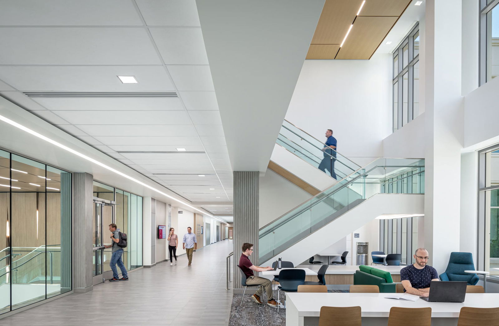 A vibrant collaborative space in the Miami University Health Sciences Building featuring a two-story atrium with modern design elements. The area includes multiple seating arrangements with wooden, black plastic, and blue fabric chairs arranged around white tables where two students work independently. A central white staircase with glass railings connects to upper levels where people are visible in motion.