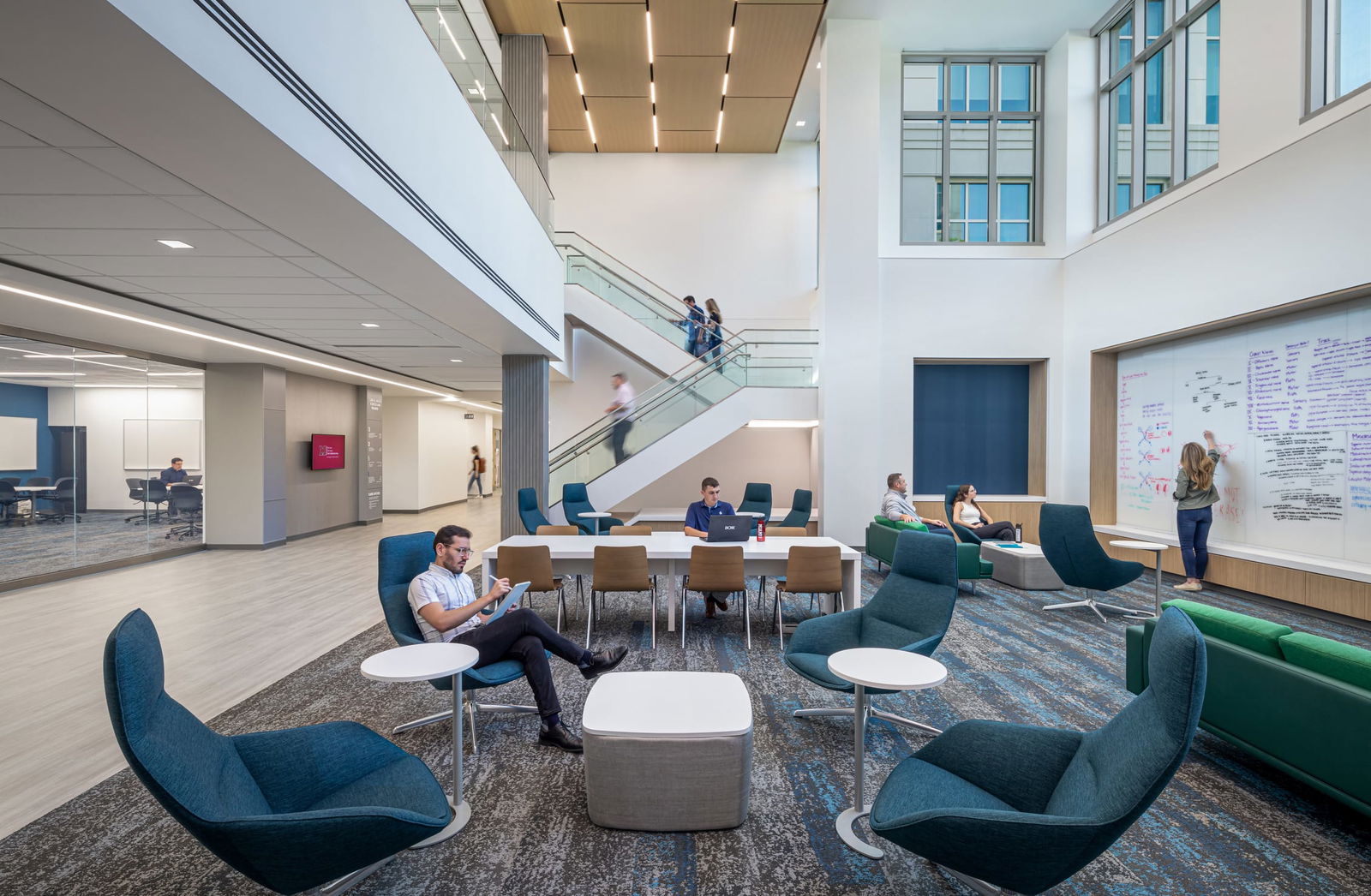 A vibrant collaborative space in the Miami University Health Sciences Building featuring a two-story atrium with modern design elements. The area includes multiple seating arrangements with blue and green furniture on patterned carpeting, where students work independently and in groups. A central white staircase with glass railings connects to upper levels where people are visible in motion. The space incorporates areas for various activities, including a whiteboard collaboration wall where someone is writing, comfortable lounge seating, and formal meeting tables. Large windows allow natural light to flood the space, complementing the warm wood accents and clean white surfaces that create a professional yet inviting learning environment.