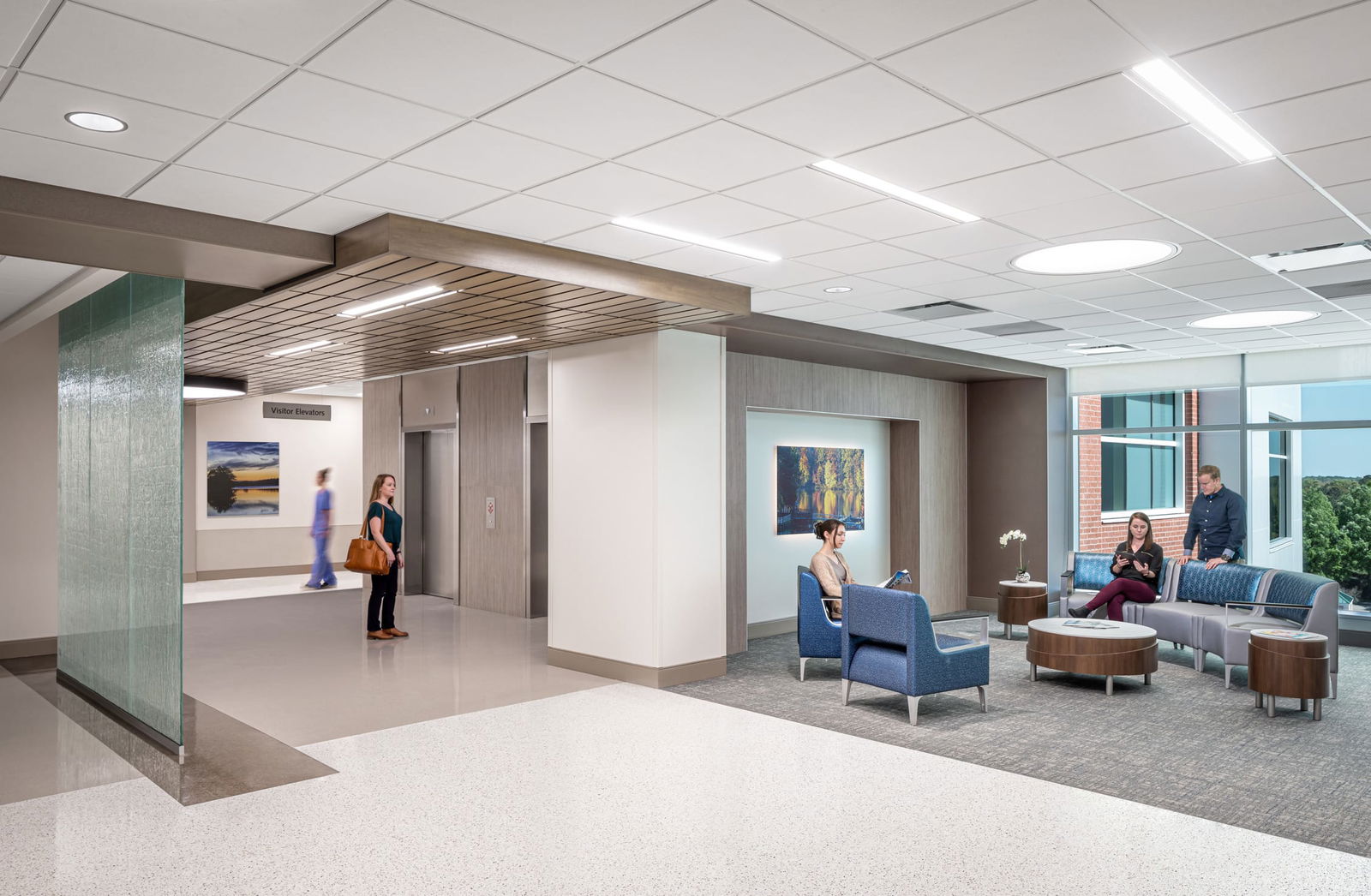 Modern medical facility interior with visitor elevator area and waiting lounge. A glass-walled entrance leads to a seating area with blue and gray chairs, wooden round tables, and large windows overlooking green trees. Two people are waiting near the elevators, and three people are seated in the lounge area, with artwork hanging on the walls.