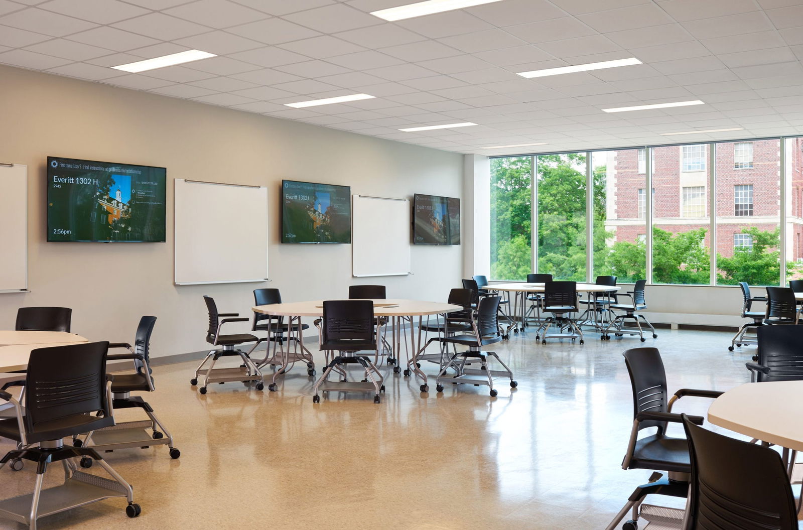 Modern classroom or collaborative workspace in Everitt Laboratory featuring round tables with black office chairs on wheels arranged throughout a bright space with polished floors. Multiple display screens and whiteboards line the walls, and large windows overlook green trees and a brick building exterior. The room has recessed lighting and a clean, contemporary educational environment design.