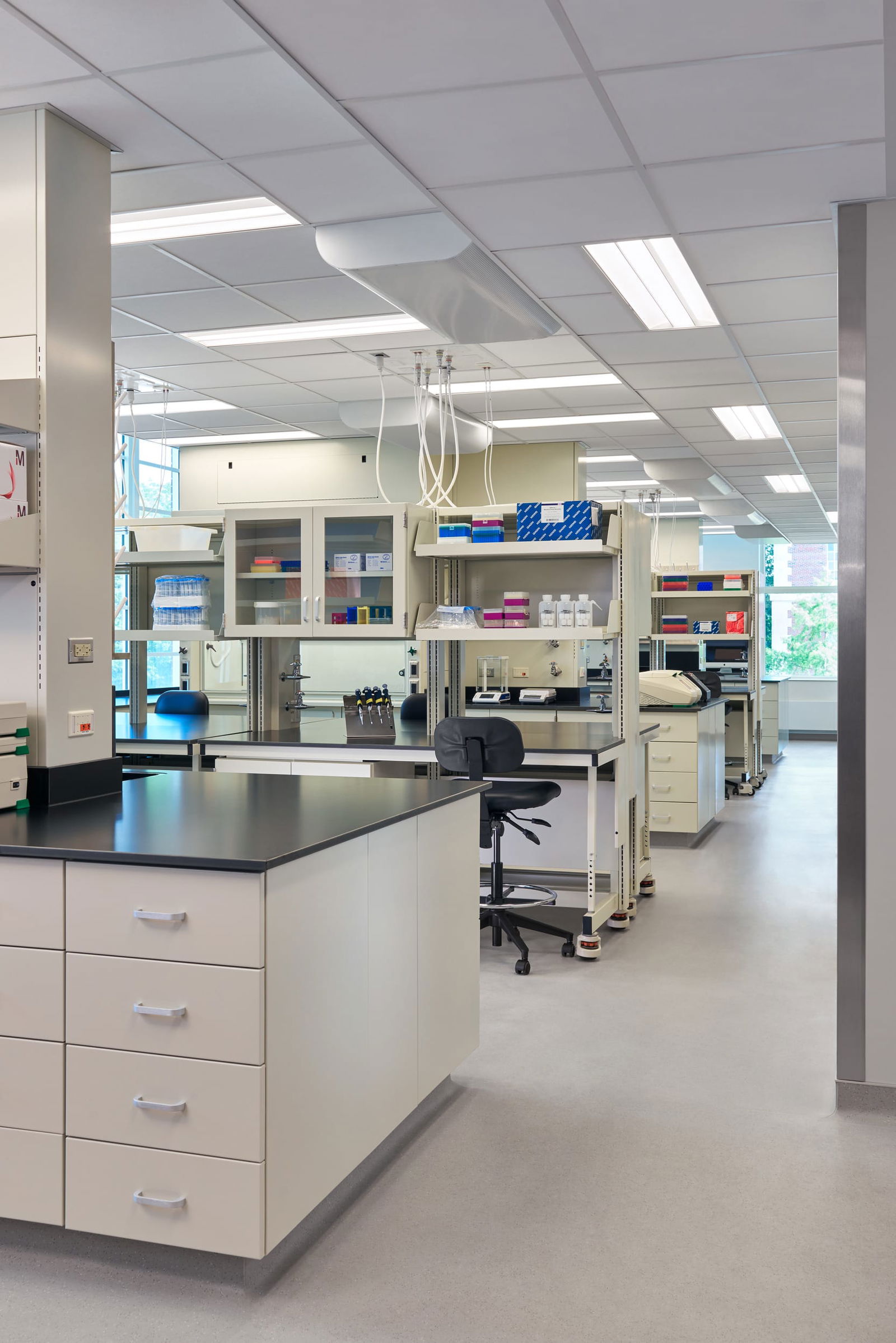 A well-equipped scientific laboratory in Everitt Laboratory with black countertops, white storage cabinets, and organized workstations. The image shows overhead utility connections, glass-fronted storage cabinets containing colorful lab supplies, and rolling chairs at workbenches. Natural light enters through windows in the background, while the foreground features drawer units and clean work surfaces designed for conducting experiments and research in a controlled environment.