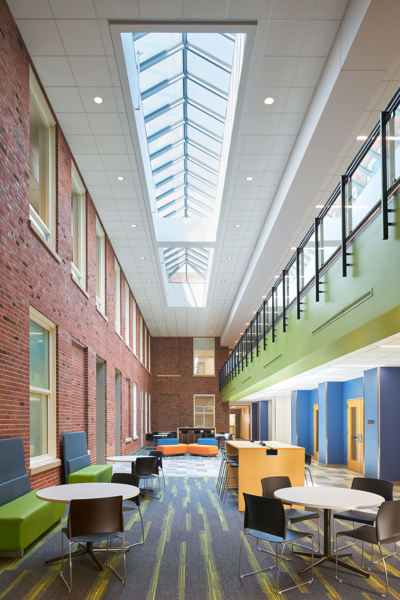 An airy, modern atrium inside Everitt Laboratory featuring a glass skylight that runs the length of the ceiling, allowing natural light to flood the space. The interior combines exposed brick walls with contemporary design elements, including colorful seating areas with round tables and lime green accents. The two-story space showcases blue and yellow wall segments, patterned carpeting, and an open second-floor balcony with metal railings, creating an inviting collaborative environment for students and faculty.