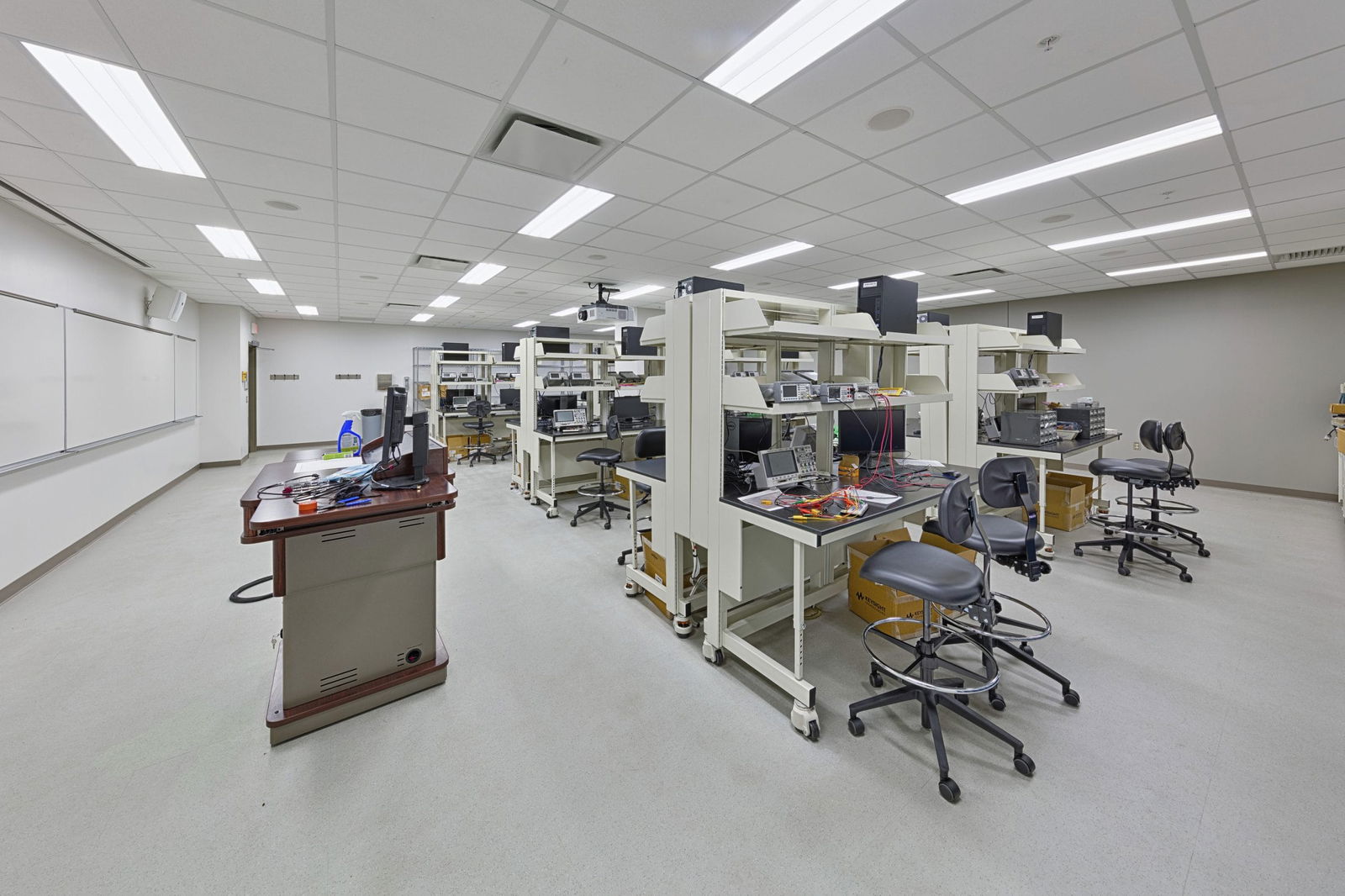 A modern engineering laboratory in Everitt Laboratory with multiple workstations featuring black adjustable stools arranged along benches designed or two people and equipped with various testing equipment and computers. The spacious room includes three side-by-side white boards with an instructor desk right in front of them. The bright, well-lit space is designed for hands-on technical education with ample workspace for students conducting experiments and projects.