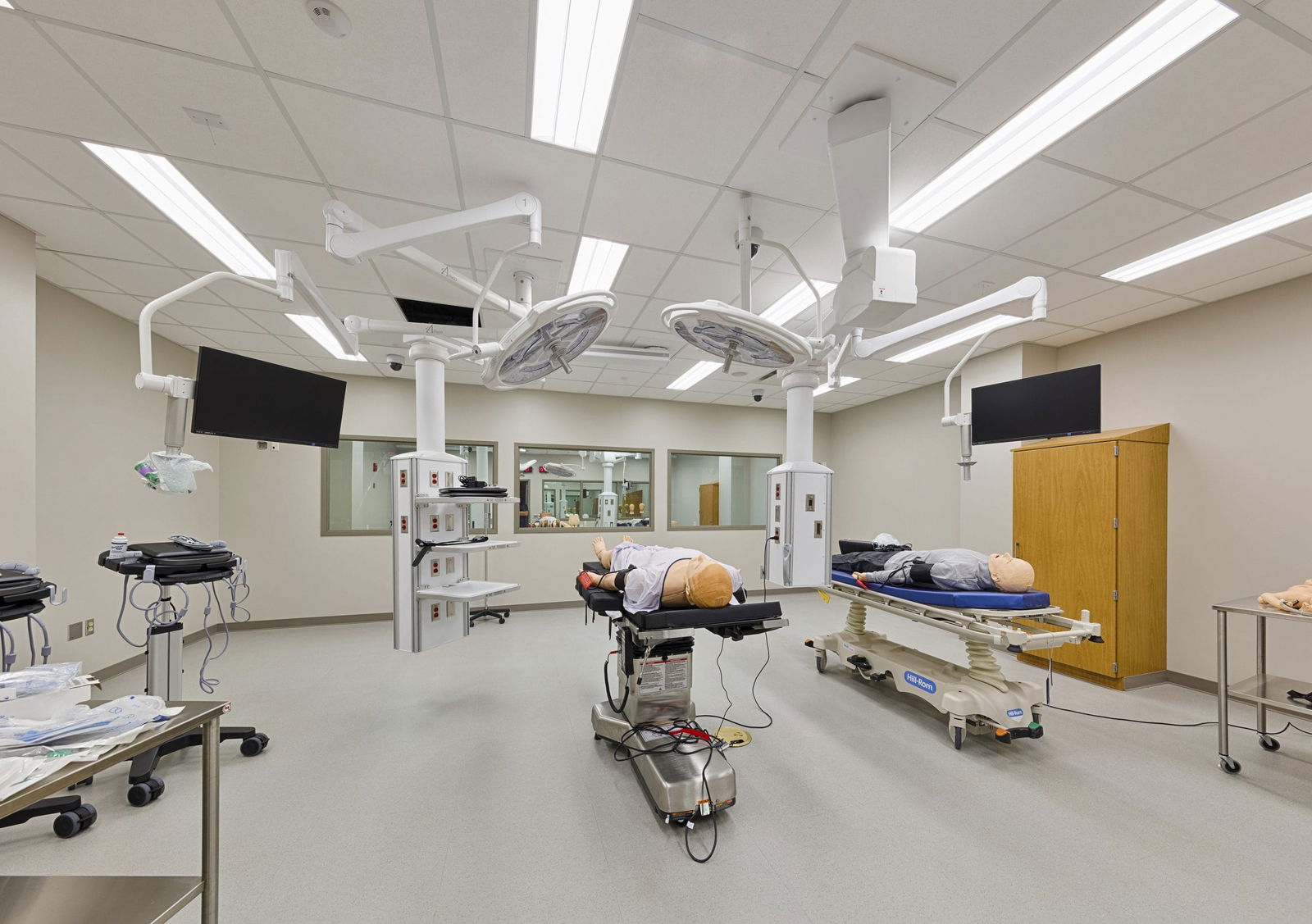 A medical simulation lab in Everitt Laboratory featuring two medical training mannequins on exam tables positioned beneath ceiling-mounted surgical lights and equipment. The clinical space includes overhead monitors, viewing windows along one wall for observation, and medical equipment on side tables. The clean, well-lit environment with gray floors and neutral walls is designed to replicate a realistic healthcare setting for educational purposes.