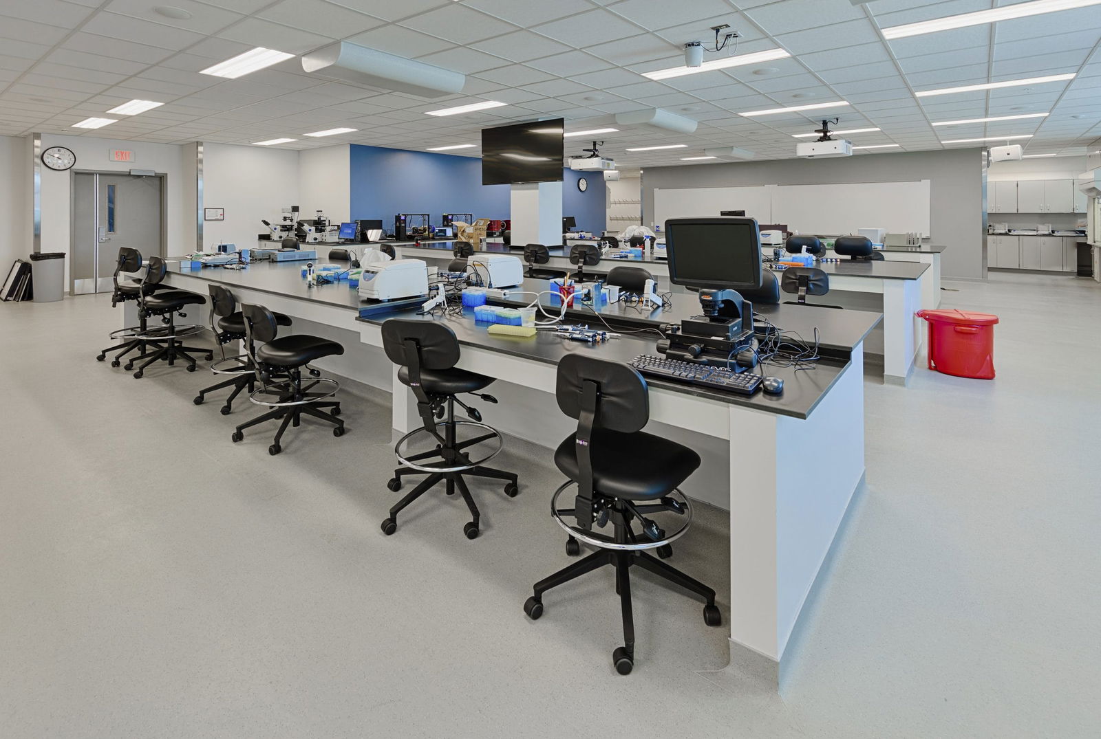 A modern engineering laboratory in Everitt Laboratory with multiple workstations featuring black adjustable stools arranged along white lab benches equipped with various testing equipment and computers. The spacious room includes ceiling-mounted monitors and projectors, a blue accent wall, and a sink area visible in the background. The bright, well-lit space is designed for hands-on technical education with ample workspace for students conducting experiments and projects.