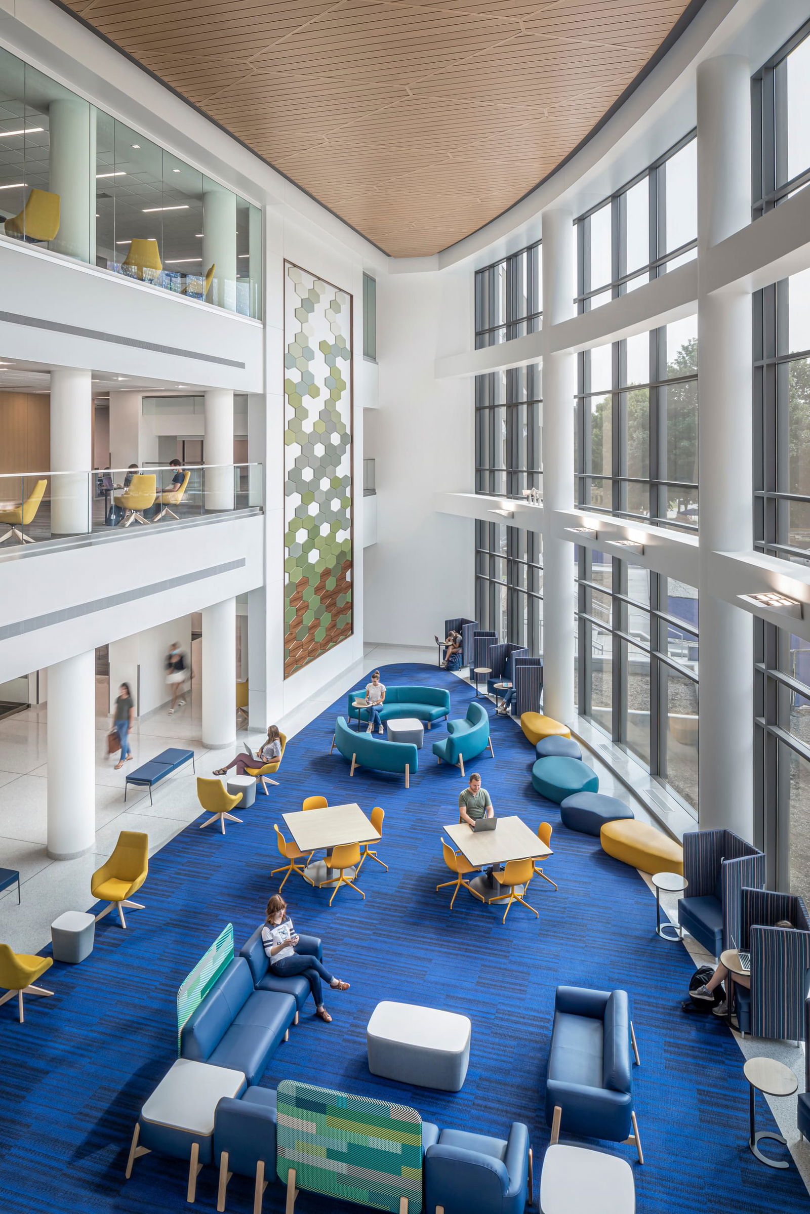 A view from the third floor overlooking a spacious, modern atrium at Butler University's Sciences Center featuring floor-to-ceiling windows that allow for ample natural light. The space includes various seating options, such as yellow and blue chairs, lounge furniture, and blue, teal, and yellow ottomans. Students are engaged in study and conversation. The interior design incorporates blue carpeting, white columns, and wood-accented ceilings, creating a bright and inviting atmosphere for learning and collaboration.