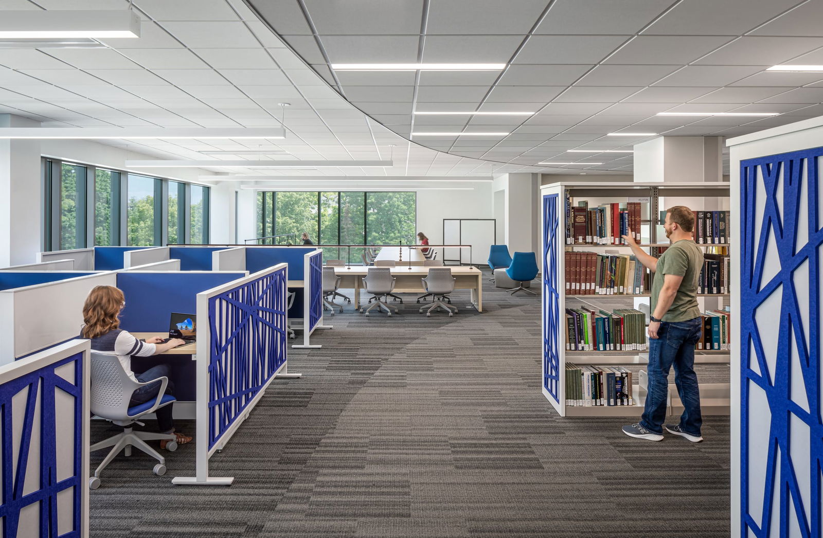 This image shows a modern academic space within the Butler University Sciences Complex with multiple functional areas. On the left side, there are cubicle-style workstations with blue dividers where a person with long hair is working at a computer. In the center background, there's a collaborative table area with white chairs where people appear to be studying near large windows that provide natural light and views of trees outside. On the right side, bookshelves are visible with a person browsing through books. The space features distinctive blue decorative panels with geometric patterns that serve as room dividers. The room has a dropped ceiling with recessed lighting and gray carpeted flooring throughout. The overall design combines open collaborative spaces with more private work areas in a contemporary academic environment.