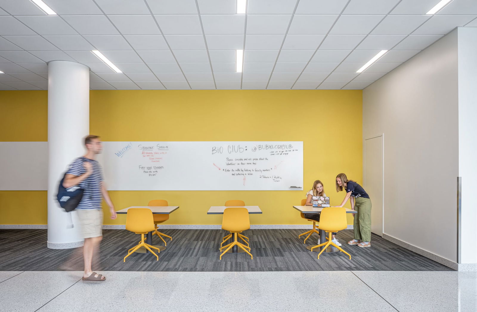 The image shows a modern collaborative study area with bright yellow walls. A large whiteboard on the wall displays notes about a