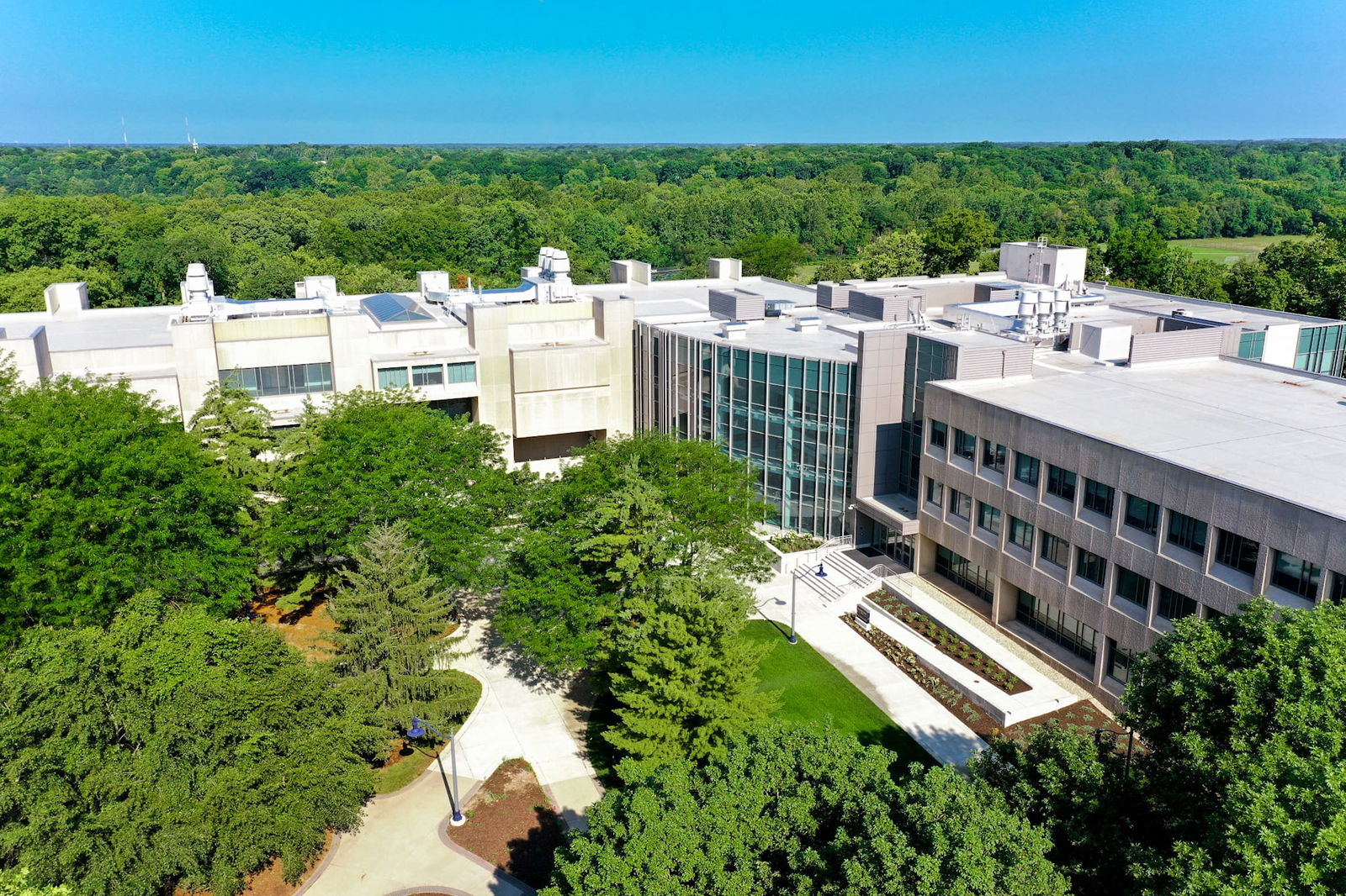This image shows the exterior of the Butler Sciences Complex from the view of a distant drone. The architecture features a blend of contemporary design elements, with a prominent glass curtain wall in the center section that creates a transparent atrium or entrance area. This glass façade reflects the blue sky and surrounding environment. The building has a beige/tan concrete or stone exterior on either side of the glass section, with regularly spaced windows. There's a formal entrance with white stairs and metal railings leading up to the building. The building is surrounded on all sides by dense, green trees and these dense trees continue all the way to the horizon line.