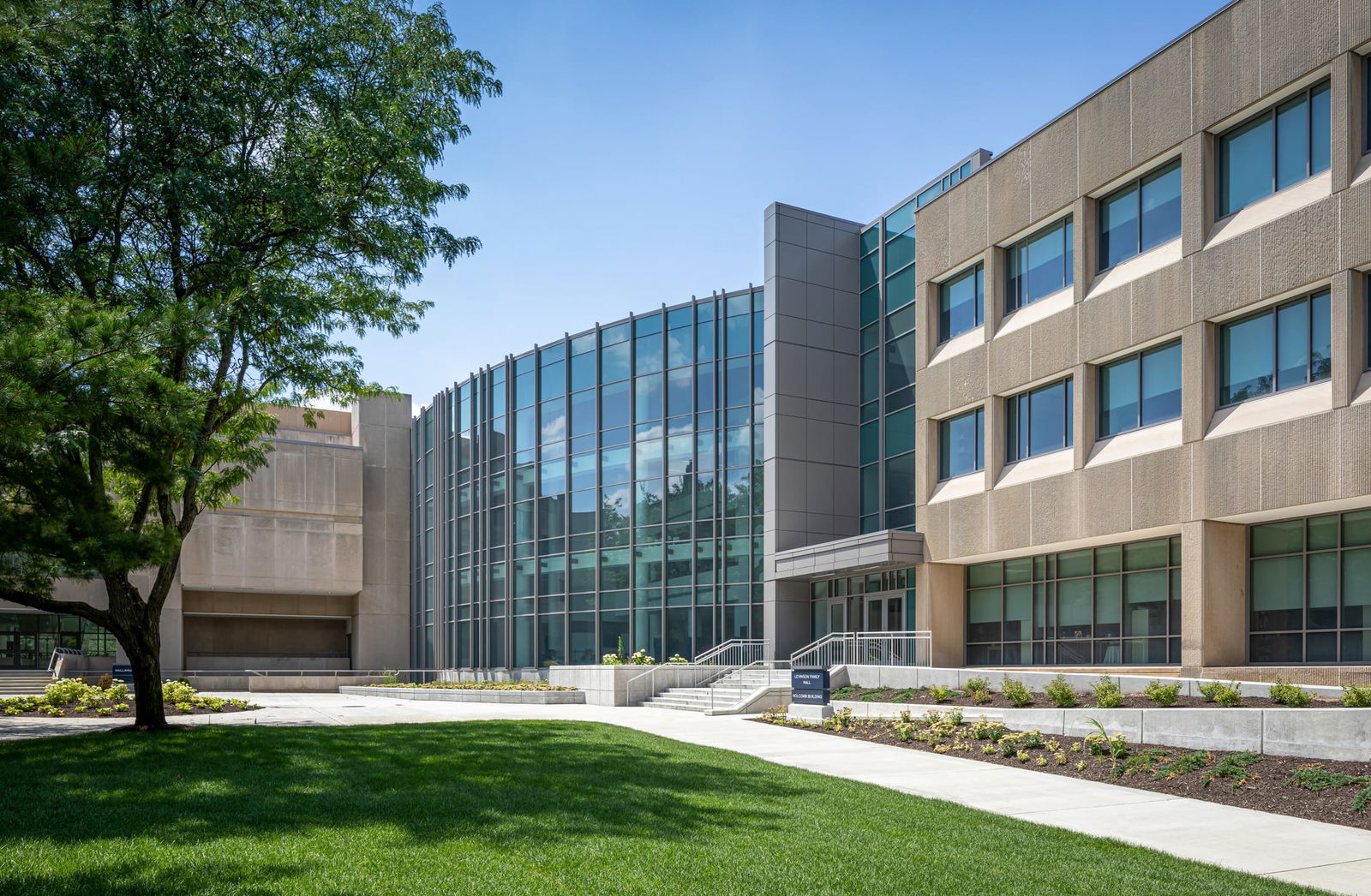 This image shows the exterior of the Butler Sciences Complex. The architecture features a blend of contemporary design elements, with a prominent glass curtain wall in the center section that creates a transparent atrium or entrance area. This glass façade reflects the blue sky and surrounding environment. The building has a beige/tan concrete or stone exterior on either side of the glass section, with regularly spaced windows. There's a formal entrance with white stairs and metal railings leading up to the building. The landscaping around the building is well-maintained, with a manicured lawn in the foreground, freshly planted garden beds with young plants near the entrance, and a mature shade tree on the left side providing some natural contrast to the architectural elements.