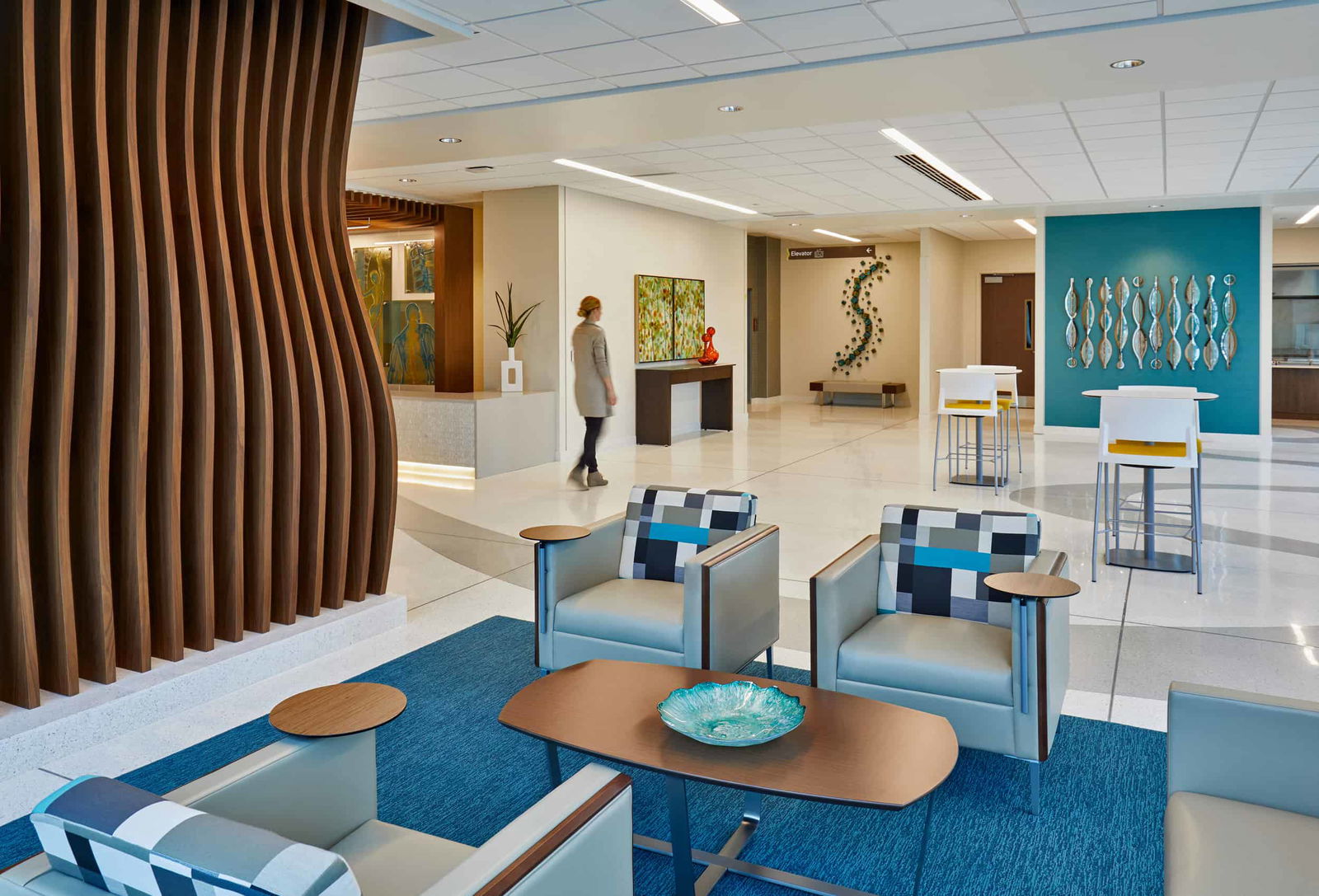 The lobby is filled with contemporary elements, including modern blue and grey furniture with warm wood features. The healthcare interior design incorporates artwork and organic forms, creating a soothing environment. Sculptures and custom-designed woodwork reflect the curves of the spine, highlighting the Spine Hospital’s specialized focus. This image shows BSA's expertise in interior design.