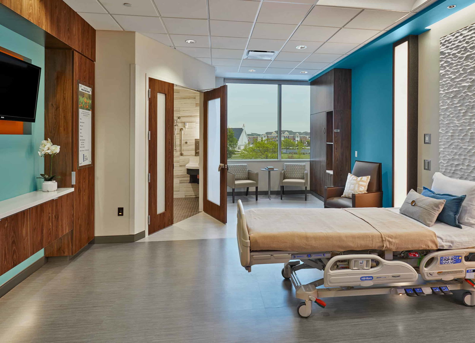 This image shows a luxury-feeling patient room for overnight hospital stays, but this is the standard room for the Indiana Spine Hospital. Warm wooden accents in the doors, furniture, and bed contribute to the luxury feel. The open bathroom doors show a spa-like luxury bathroom with floor-to-ceiling tile and a standing shower for enhanced patient safety. This image shows BSA's expertise in interior design, specifically our expertise in designing patient-centered spaces for patients who have just undergone an operation to the spine.