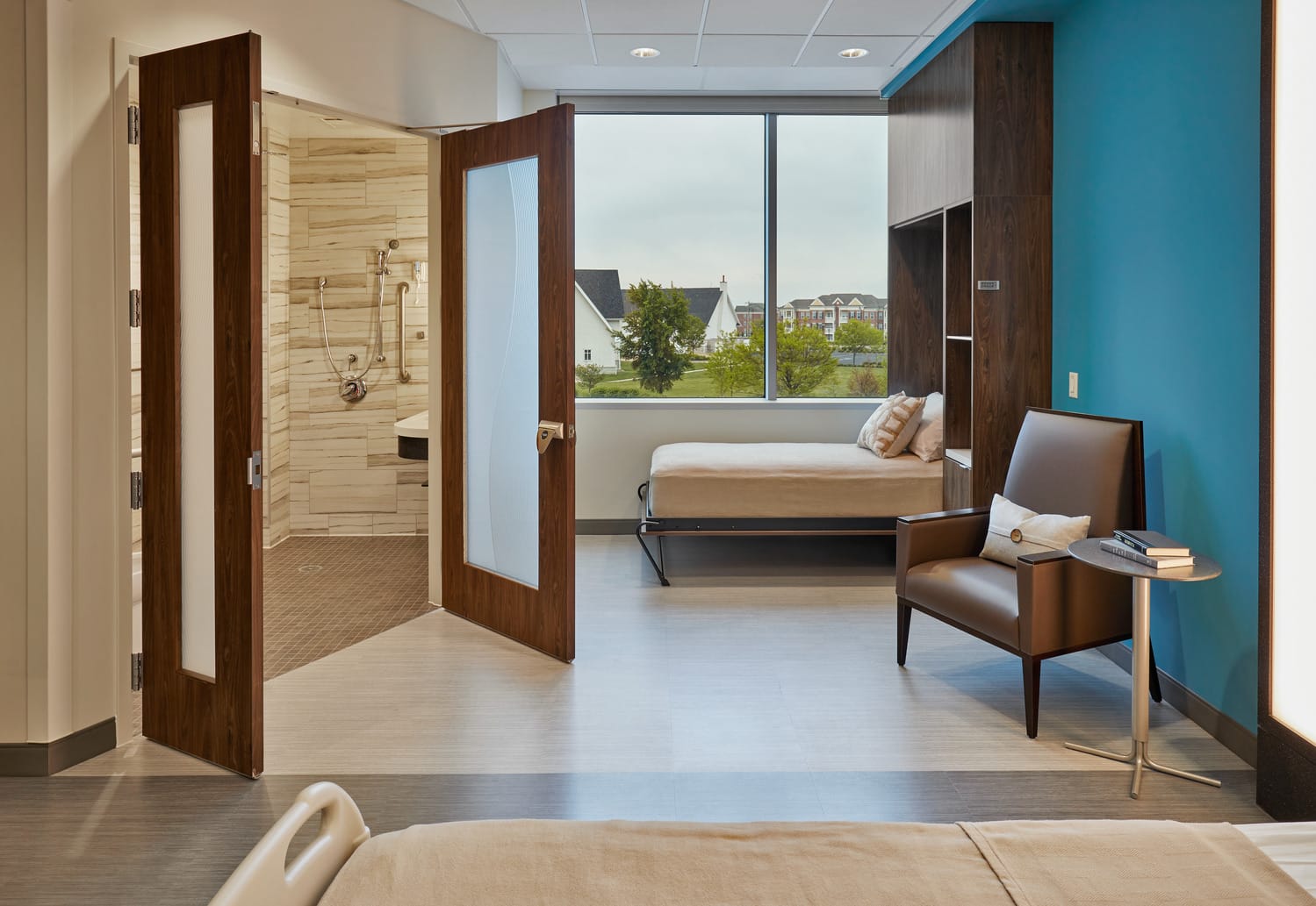 This image shows a luxury-feeling patient room for overnight hospital stays, but this is the standard room for the Indiana Spine Hospital. Warm wooden accents in the doors, furniture, and bed contribute to the luxury feel. The bed looks more like a residential bed than a hospital bed, contributing to a hospitality-like feel. The open bathroom doors show a spa-like luxury bathroom with floor-to-ceiling tile and a standing shower for enhanced patient safety. This image shows BSA's expertise in interior design, specifically our expertise in designing patient-centered spaces for patients who have just undergone an operation to the spine.