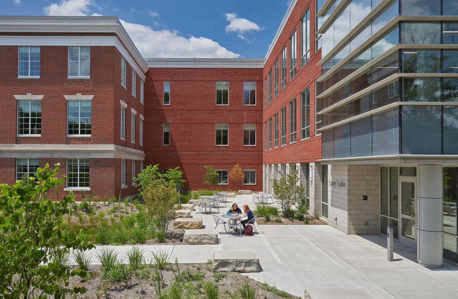 This image shows a courtyard area between sections of the Barnes Science Hall at Franklin College. The structure features traditional red brick architecture with a modern glass section visible on the right side. The courtyard is designed as an outdoor gathering space with concrete paving, several metal tables and chairs where two people can be seen sitting and conversing. The space includes new landscaping with young shrubs, ornamental grasses, and stone elements integrated into the design. The courtyard is surrounded on three sides by the brick building, creating a semi-enclosed outdoor area. The entrance to one section of the building is visible on the right, with what appears to be signage near the doorway. The scene is captured on a bright day with blue skies and some clouds, highlighting the contrast between the traditional red brick sections and the contemporary glass façade.