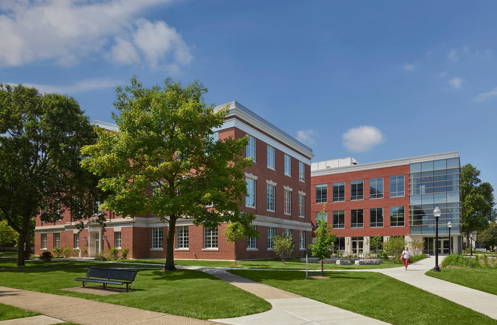 The image shows Barnes Science Hall at Franklin College. The building features traditional red brick architecture with a combination of classic and modern design elements. The structure appears to have multiple wings, with a more traditional wing on the left featuring regular windows with white trim and a more contemporary glass-fronted section on the right. The building is set on a well-maintained campus green with manicured lawns, mature trees providing shade, and concrete walkways leading to the entrances. There's a bench visible in the foreground, and a person can be seen walking on one of the paths. Decorative lampposts are positioned along the walkways. The setting is bright and sunny with a blue sky and a few scattered clouds, creating an inviting academic atmosphere. The landscaping around the science building includes small shrubs and green spaces that enhance the collegiate environment.