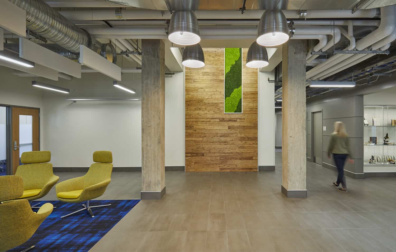 A secondary view of the contemporary collaborative space in Barnes Science Hall featuring a modern industrial design with exposed ceiling elements and hanging silver pendant lights. The area includes wooden wall panels accented by vibrant vertical moss installations, creating a biophilic element amid concrete columns. Bright blue accent walls add color to the light wood flooring, while glass railings lead to different levels. Bar-height seating is visible on the left, and a person walks down a corridor in the distance, highlighting the open, flowing layout designed for interaction and study.