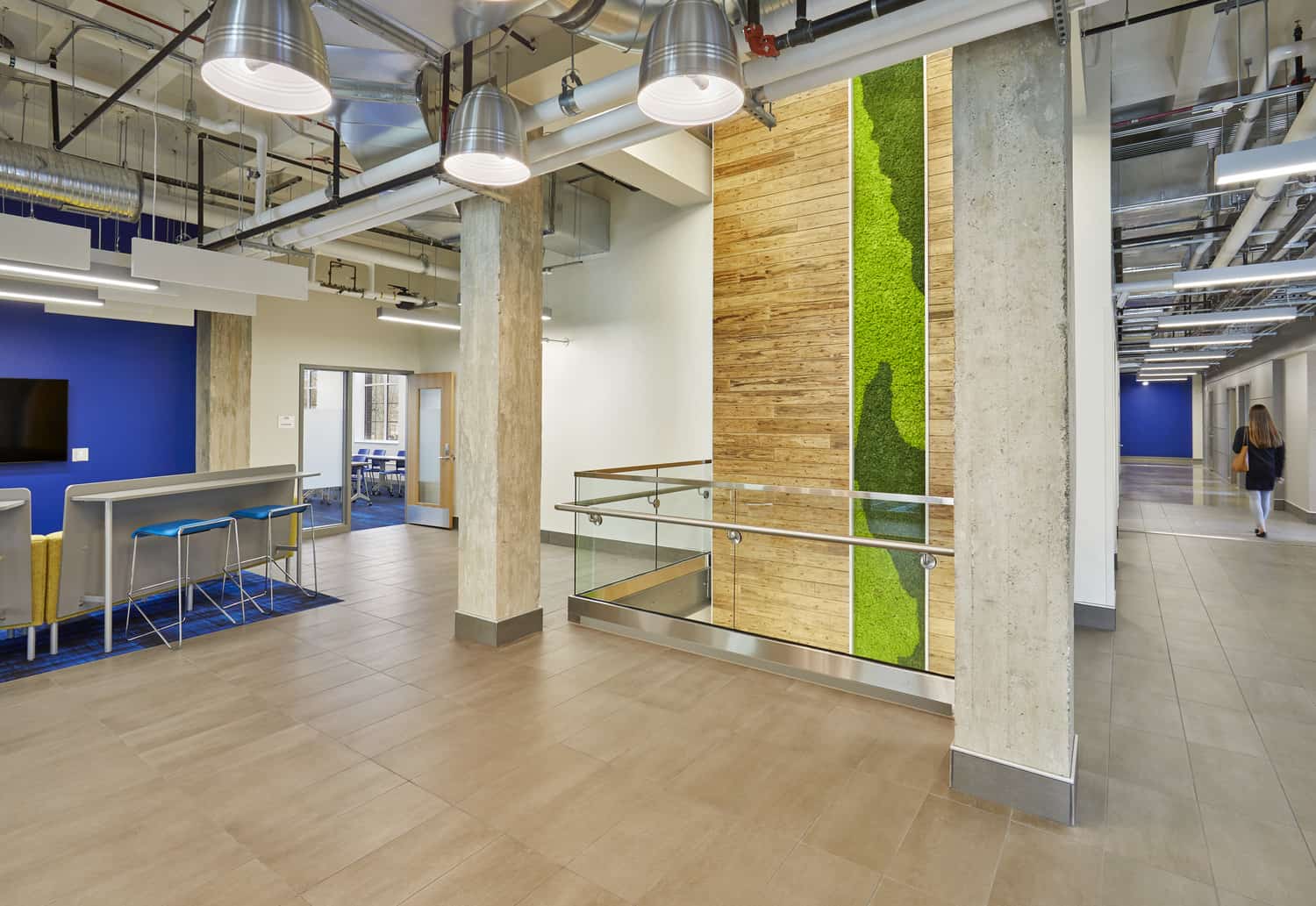 A contemporary collaborative space in Barnes Science Hall featuring a modern industrial design with exposed ceiling elements and hanging silver pendant lights. The area includes wooden wall panels accented by vibrant vertical moss installations, creating a biophilic element amid concrete columns. Bright blue accent walls add color to the light wood flooring, while glass railings lead to different levels. Bar-height seating is visible on the left, and a person walks down a corridor in the distance, highlighting the open, flowing layout designed for interaction and study.