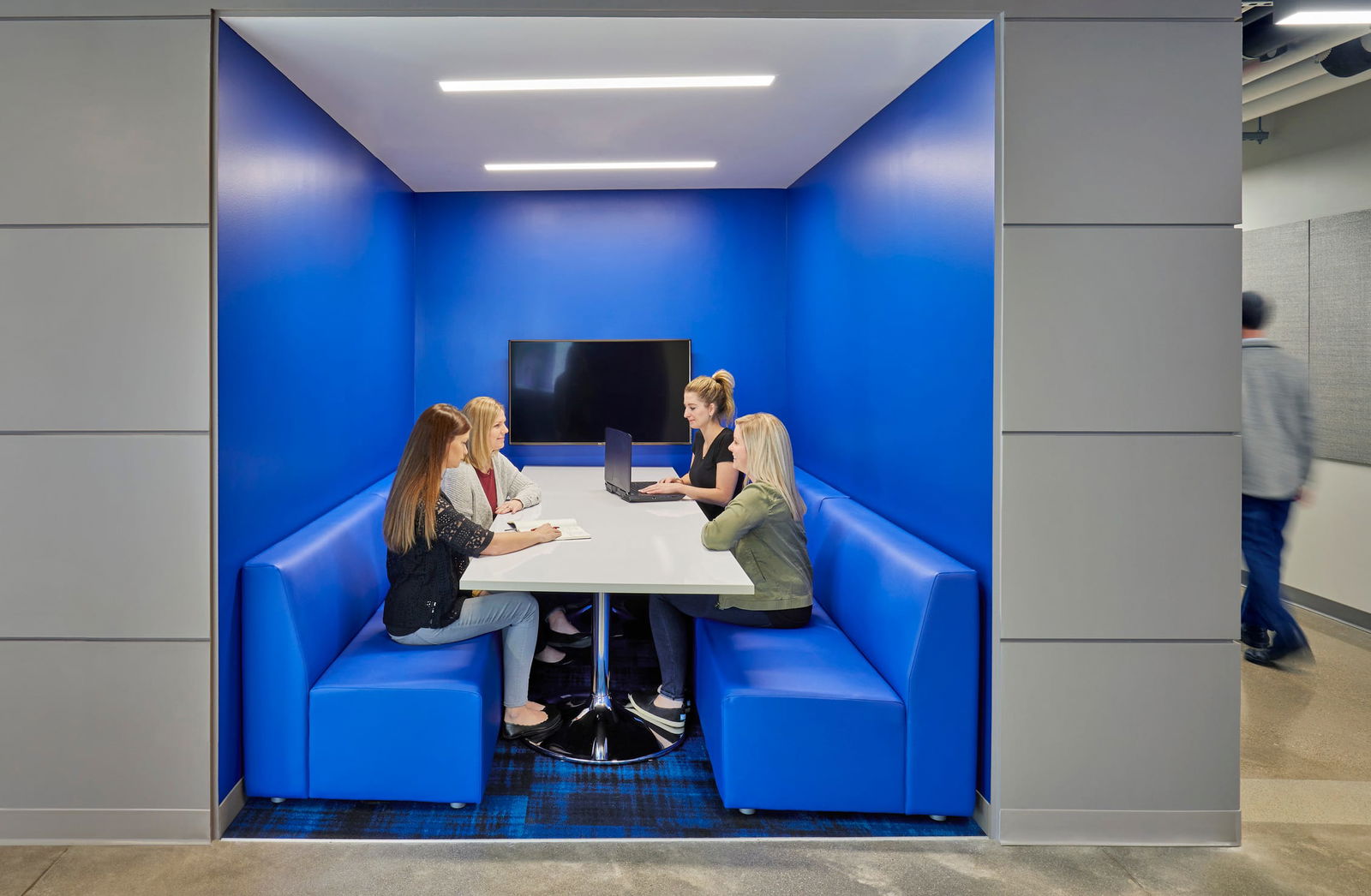 The image shows a modern collaborative workspace featuring a small meeting pod with bright blue walls and built-in booth seating. Four young women are seated around a white circular table having a discussion, with one using a laptop. The pod is designed as a semi-private alcove within what appears to be a larger office or educational space, with a flat-screen display mounted on the back wall. The blue booth seating matches the vibrant blue walls, and the space is well-lit with recessed lighting. A person can be seen walking past the pod in the hallway on the right side. The design exemplifies contemporary collaborative spaces found in modern educational facilities.