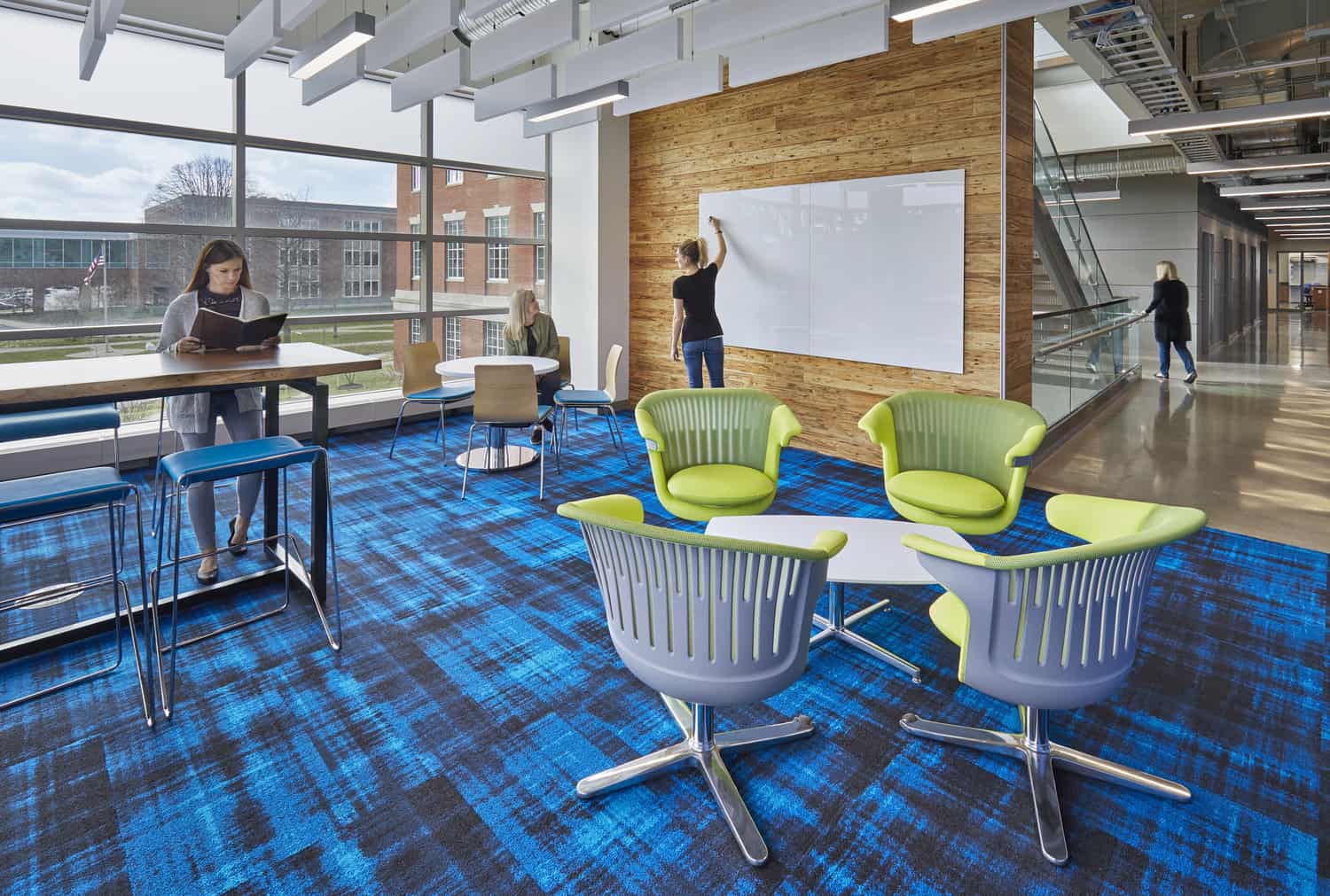 The image shows a modern educational collaborative space with vibrant blue carpeting and bright lime green chairs. A student studies at a standing-height table on the left, while another person writes on a whiteboard mounted on a wooden accent wall. Floor-to-ceiling windows provide natural light and views of brick campus buildings outside. The space features contemporary furniture including swivel chairs with plastic shells arranged around small tables. A glass-walled corridor runs along the right side where a person is walking. The design combines open study areas with presentation spaces, exemplifying contemporary learning environment design with its mix of collaborative furniture, flexible spaces, and bright accent colors.