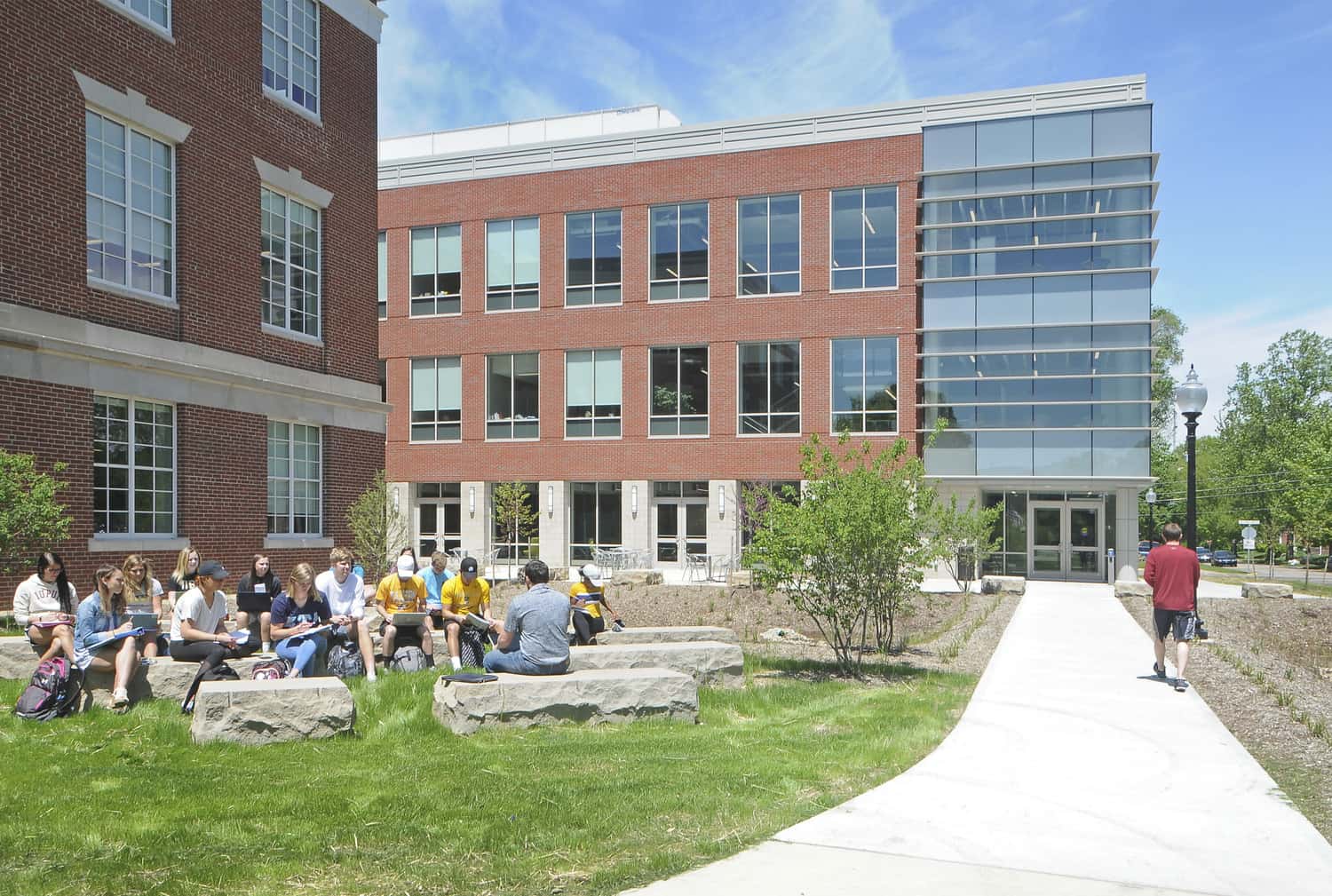 This image shows a courtyard area between sections of the Barnes Science Hall at Franklin College. The structure features traditional red brick architecture with a modern glass section visible on the right side. The courtyard is designed as an outdoor gathering space. Over a dozen students appear to be studying, sitting on large stone elements that act as benches. The scene is captured on a bright day with blue skies and some clouds, highlighting the contrast between the traditional red brick sections and the contemporary glass façade.
