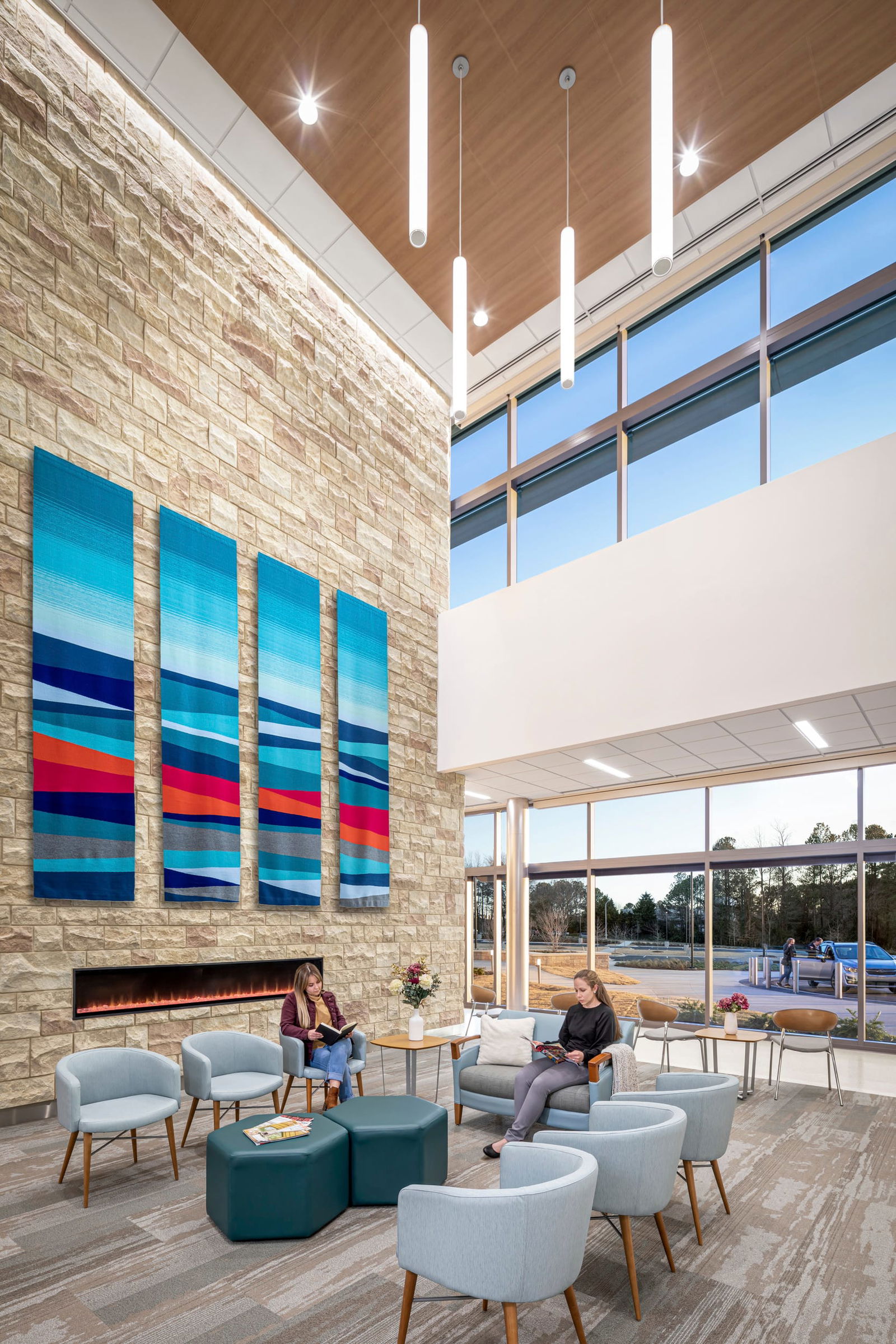 Spacious, modern interior patient waiting room of the UNC Rex Cancer Center with high ceilings and floor-to-ceiling windows. A stone accent wall features four large, colorful abstract art panels in blue and red tones. A linear fireplace is mounted below the artwork. The space is furnished with light blue and green seating including chairs, ottomans, and a sofa. Two people are seated in the lounge, one reading a book and another working on a laptop. Natural light floods the room, offering a view of trees and a parking area outside.
