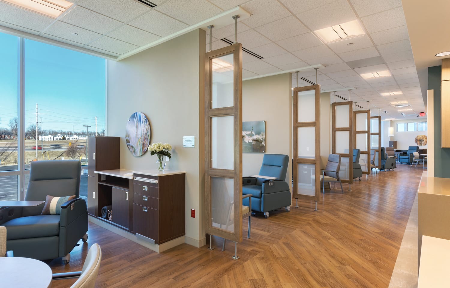 A bright and modern healthcare treatment area with semi-private patient spaces separated by wooden-framed glass dividers. The room features reclining medical chairs with armrests, warm wood flooring, and soft neutral-colored walls. Large windows allow natural light to enter, offering a view of the outdoors. Decorative artwork and plants enhance the calming atmosphere.