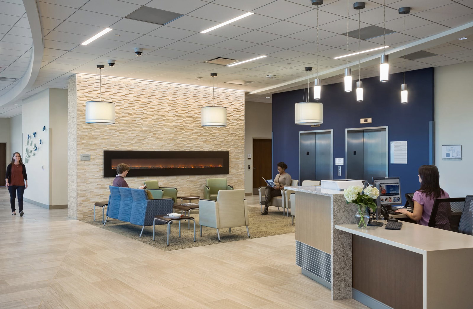 A modern hospital lobby with a reception desk, seating area, and decorative lighting. People are seated and working on laptops. Elevators are visible in the background. Green, navy blue, and brown waiting room furniture is arranged on brown rugs. This image showcases BSA's expertise in designing spaces that prioritize patient care and function.