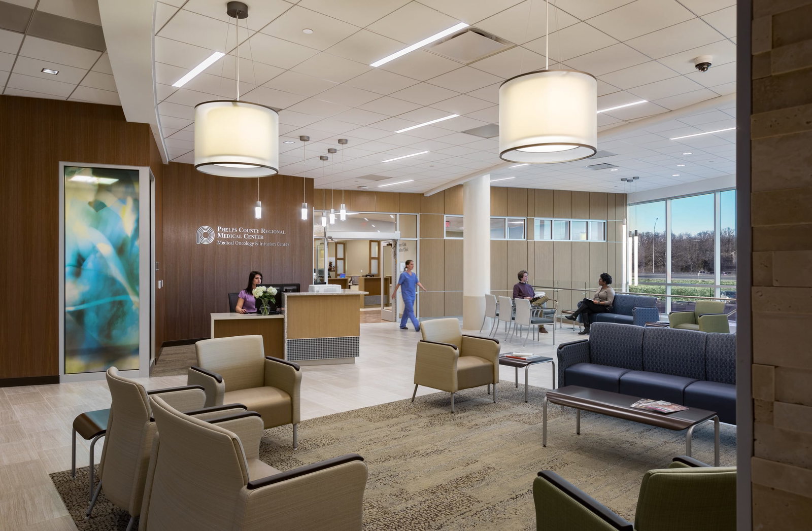 A spacious first-floor hospital lobby with sleek modern design, and large floor-to-ceiling windows allowing natural light. Neutral floor, wall colors, and walls with wood panels create a calming presence. Green, navy blue, and brown furniture is arranged on brown rugs. This image showcases BSA's expertise in designing spaces that prioritize patient care and function.