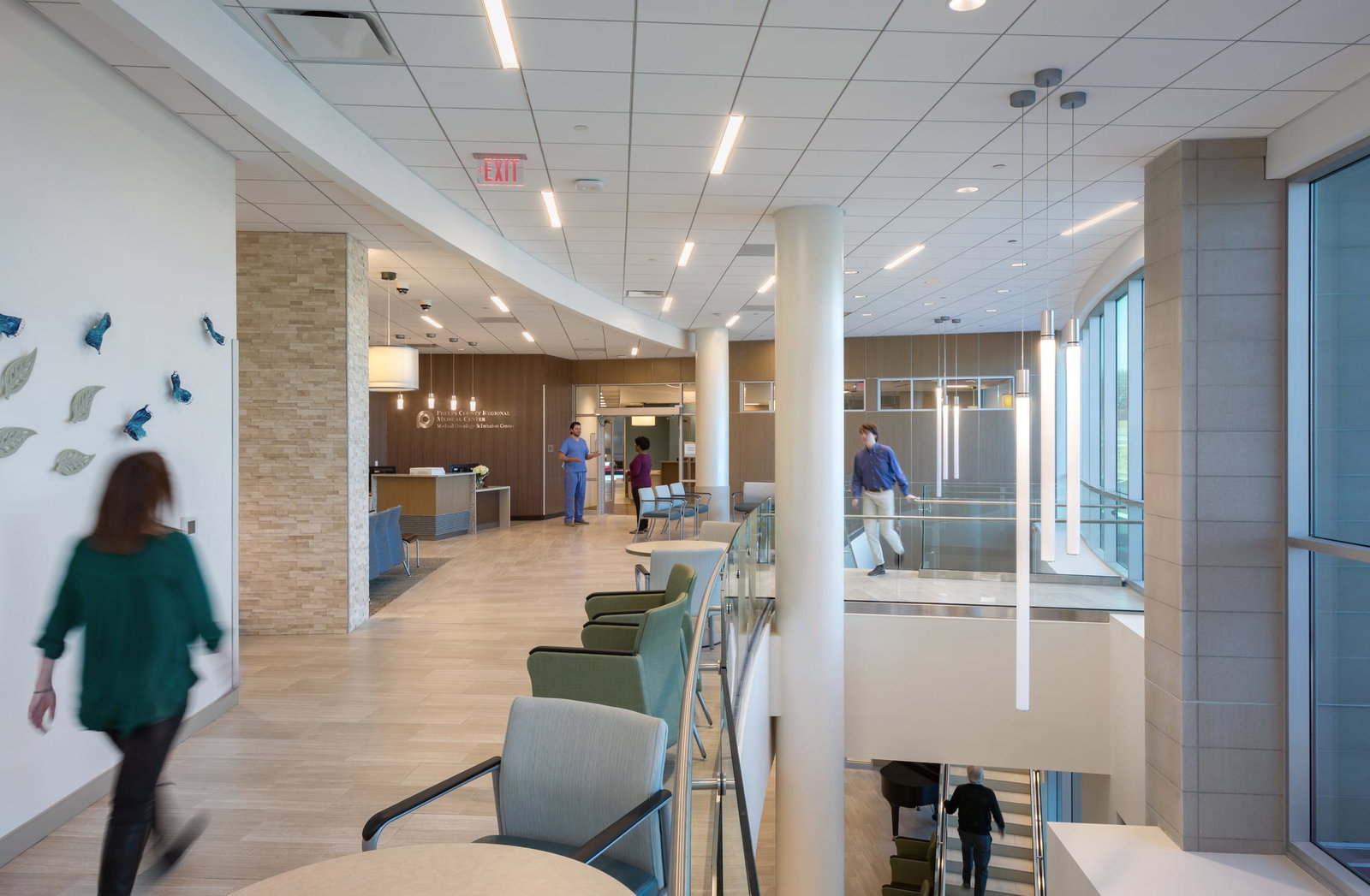 A spacious second-floor hospital lobby with high ceilings, sleek modern design, and large windows allowing natural light. Neutral floor, wall colors, and walls with decorative stone create a calming presence. Large vertical lighting fixtures hang from the ceiling, and overhead lighting lines the ceiling. This image showcases BSA's expertise in designing spaces that prioritize patient care and function.
