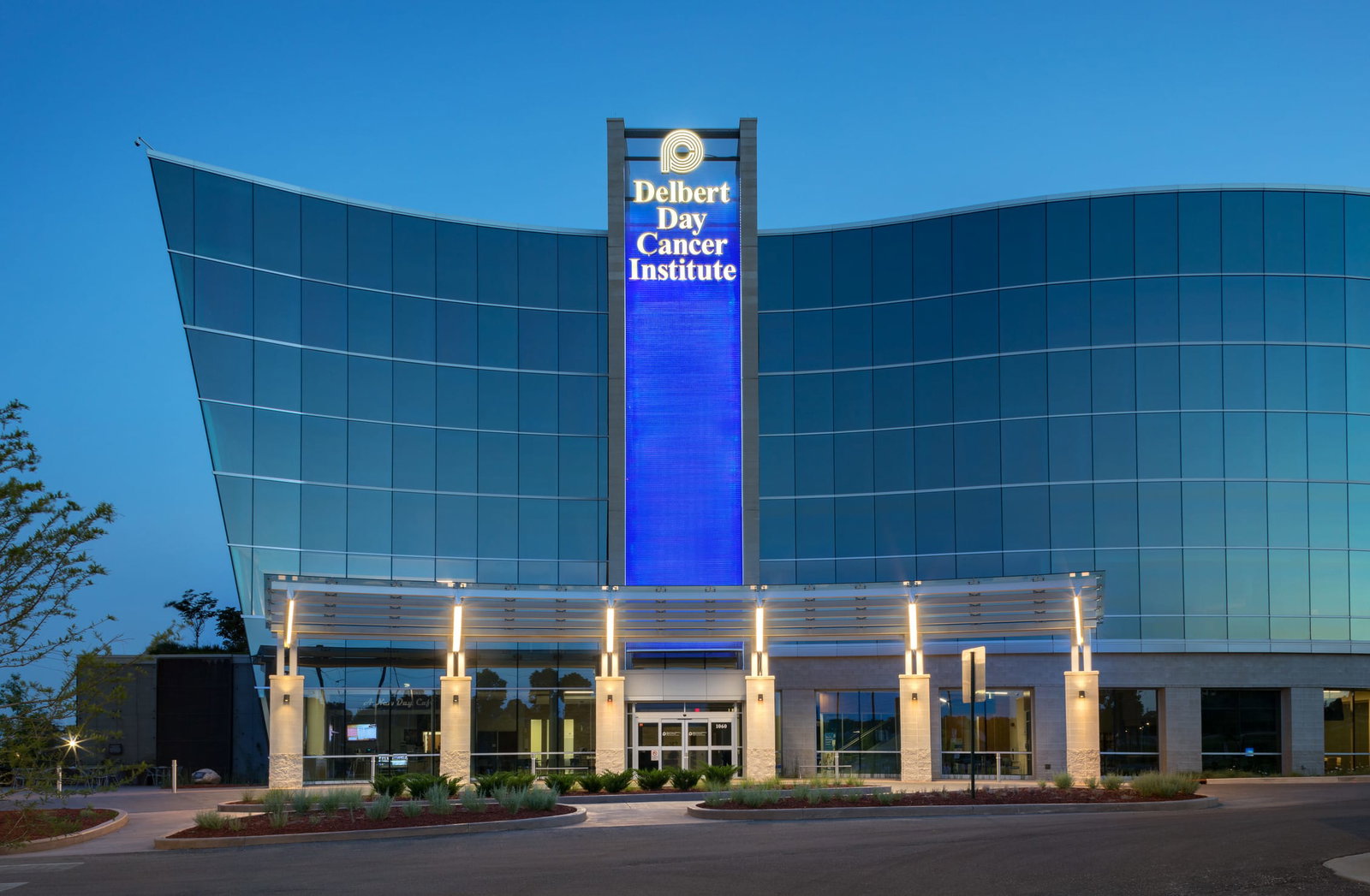 A modern glass-fronted building with a curved facade reflecting the blue sky and clouds. The structure has a central stone pillar displaying the name 'Delbert Day Cancer Institute.' The entrance features a covered driveway with a glass canopy supported by white columns. Landscaping with greenery and red mulch lines the front of the building, and a smooth driveway leads up to the main doors. This image showcases BSA's expertise in architecture and engineering.