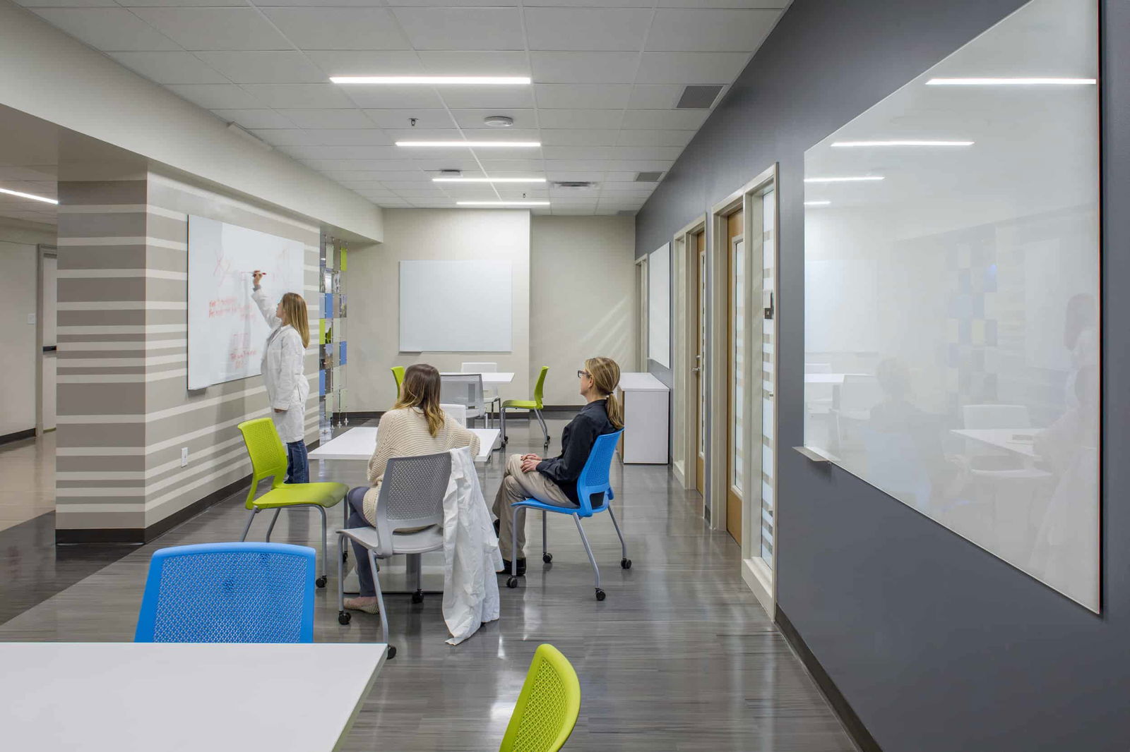 Collaborative teaching area of the UT Austin College of Natural Sciences Painter Hall featuring a modern, collaborative space. The area is not a classroom, but a casual and open instructive space with tables with bright green and blue chairs. Two women are seated and looking towards a third woman writing something on a white board on a wall with gray and white horizontal stripes. The space has a contemporary aesthetic with light-colored flooring, drop ceiling, and recessed lighting, embodying a dynamic academic environment.