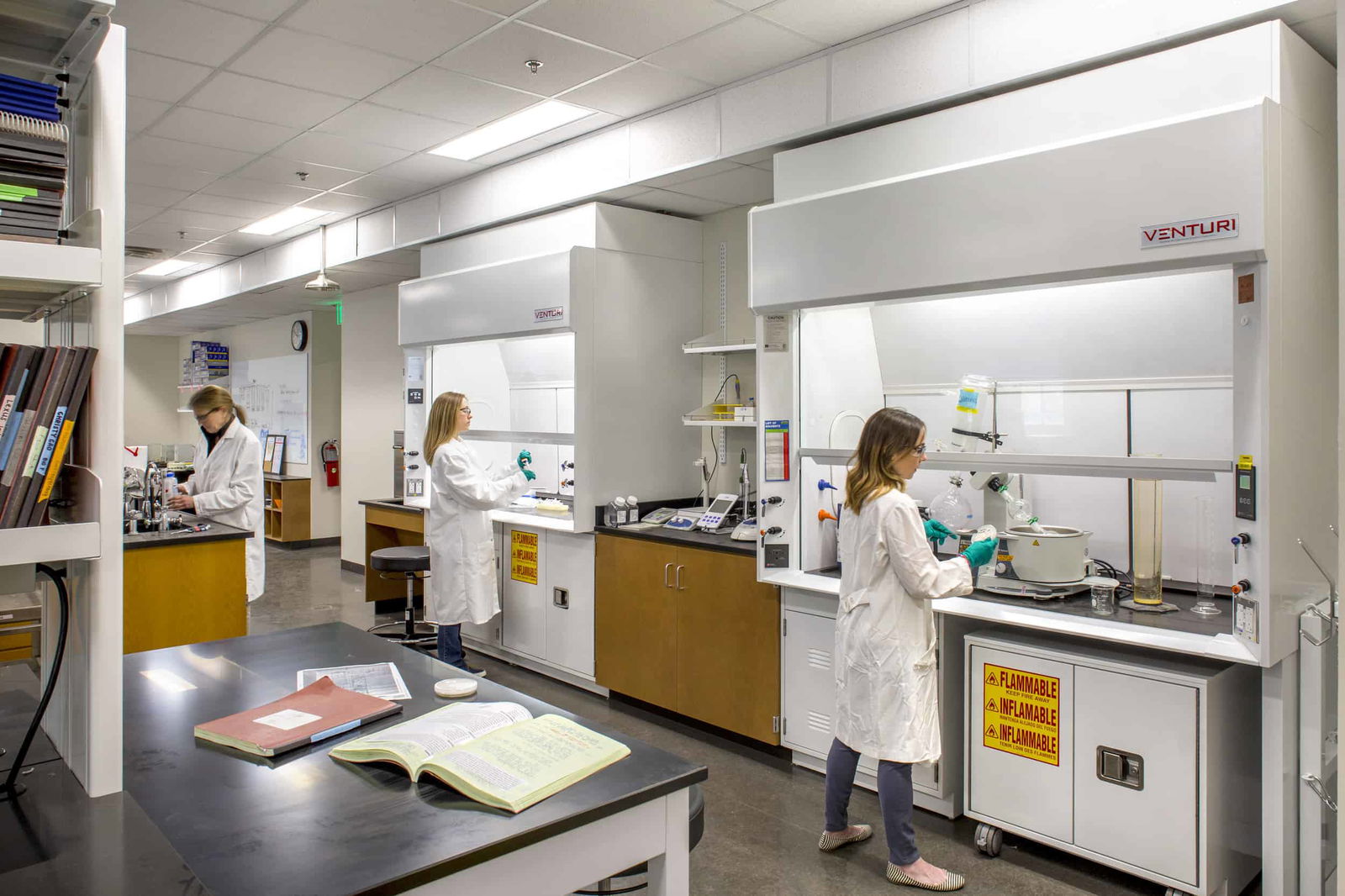 A modern scientific laboratory at UT Austin's College of Natural Sciences Painter Hall, featuring three researchers in white lab coats working at separate fume hoods labeled 'VENTURI'. The space has a professional layout with black countertops, wooden cabinetry, and white walls. In the foreground, an open reference book sits on a lab bench. Safety features are evident with flammable material storage cabinets (marked with yellow warning labels) and a fire extinguisher visible on the wall. The lab is well-lit with overhead fluorescent lighting and equipped with various scientific instruments. Bookshelves containing reference materials can be seen on the left wall, along with informational posters. The laboratory demonstrates a functional scientific workspace designed for chemical research and experimentation.
