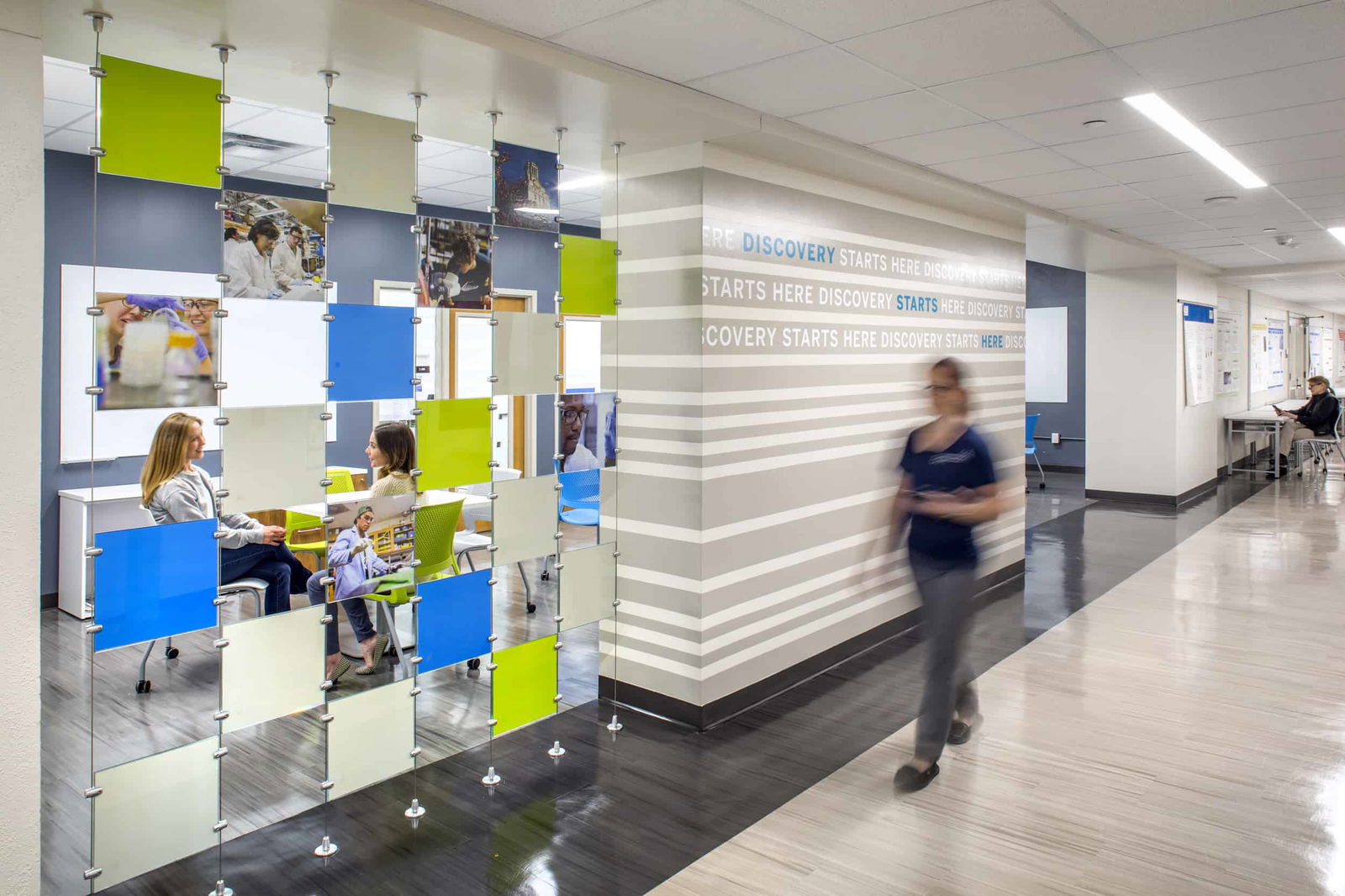 Interior corridor of the renovated UT Austin College of Natural Sciences Painter Hall featuring a modern, collaborative space. On the left, a suspended panel display system combines colorful acrylic squares in blue, lime green, and white with photographs of students in laboratory settings. Behind the panels, students are seated in a conversation area with bright green chairs. The right wall features a striking design element with gray and white horizontal stripes and the repeating text 'DISCOVERY STARTS HERE' in light blue and white. A person in motion (slightly blurred) walks through the hallway, while in the distance another person can be seen working at a desk. The space has a contemporary aesthetic with light-colored flooring, drop ceiling, and recessed lighting, embodying a dynamic academic environment.