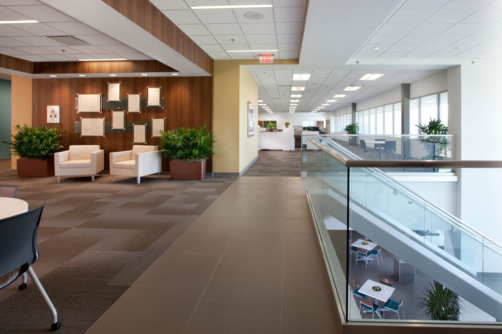 Interior hallway of the Endress+Hauser Customer Center featuring an elegant reception area with rich wood-paneled walls displaying framed documents, two white lounge chairs flanked by potted plants, and a glass railing overlooking a modern dining area below, all connected to an open workspace with abundant natural light from large windows, demonstrating the facility's contemporary design approach balancing professionalism with warmth.