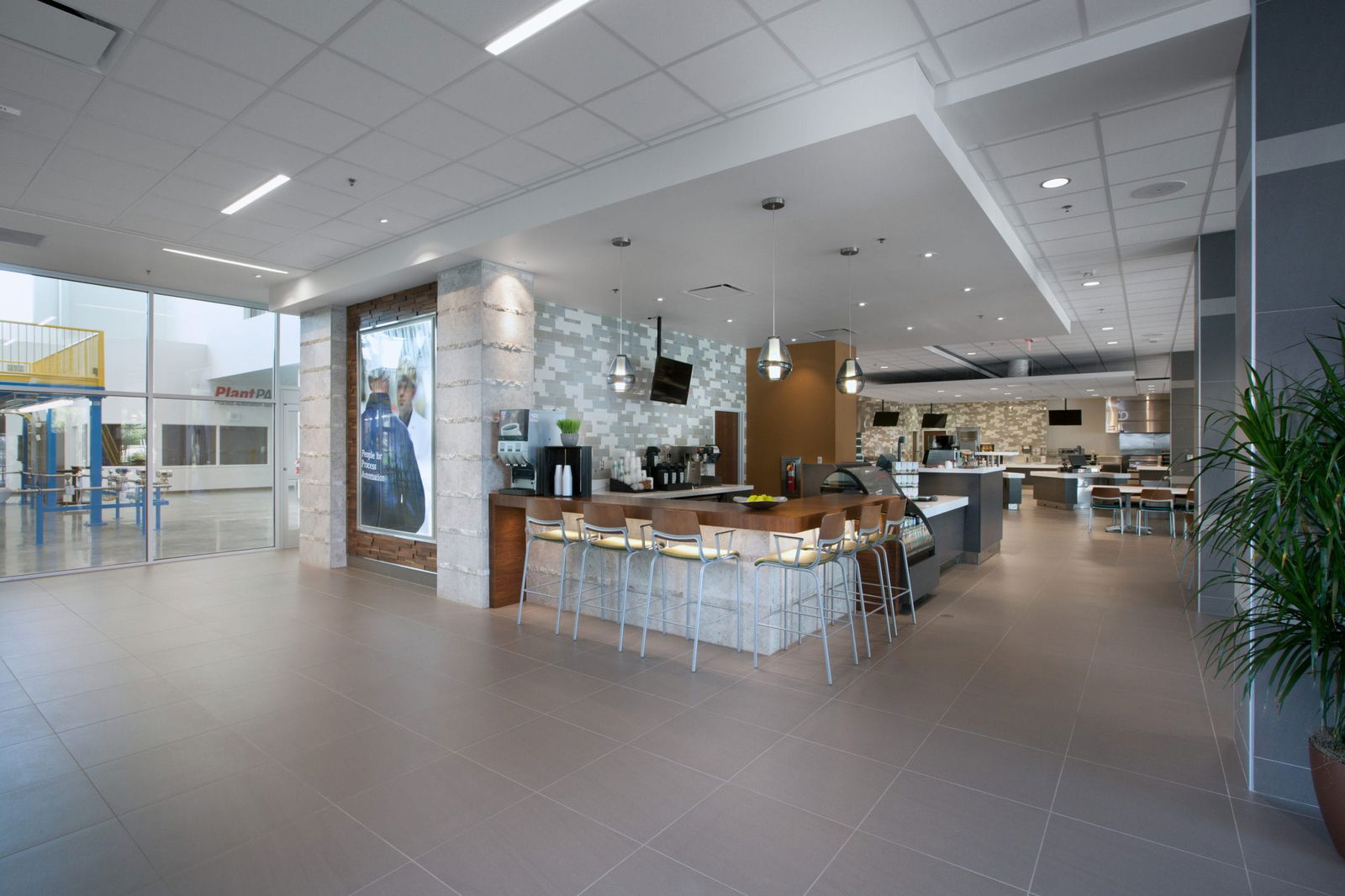 Modern, bright interior of the Endress+Hauser Customer Center featuring a stylish café area with a wooden counter, white bar stools, and coffee machines, complemented by contemporary lighting, textured wall panels, and an open layout with floor-to-ceiling windows that provide natural light and views of the exterior, all set within a professional corporate environment with sleek tile flooring and indoor plants.