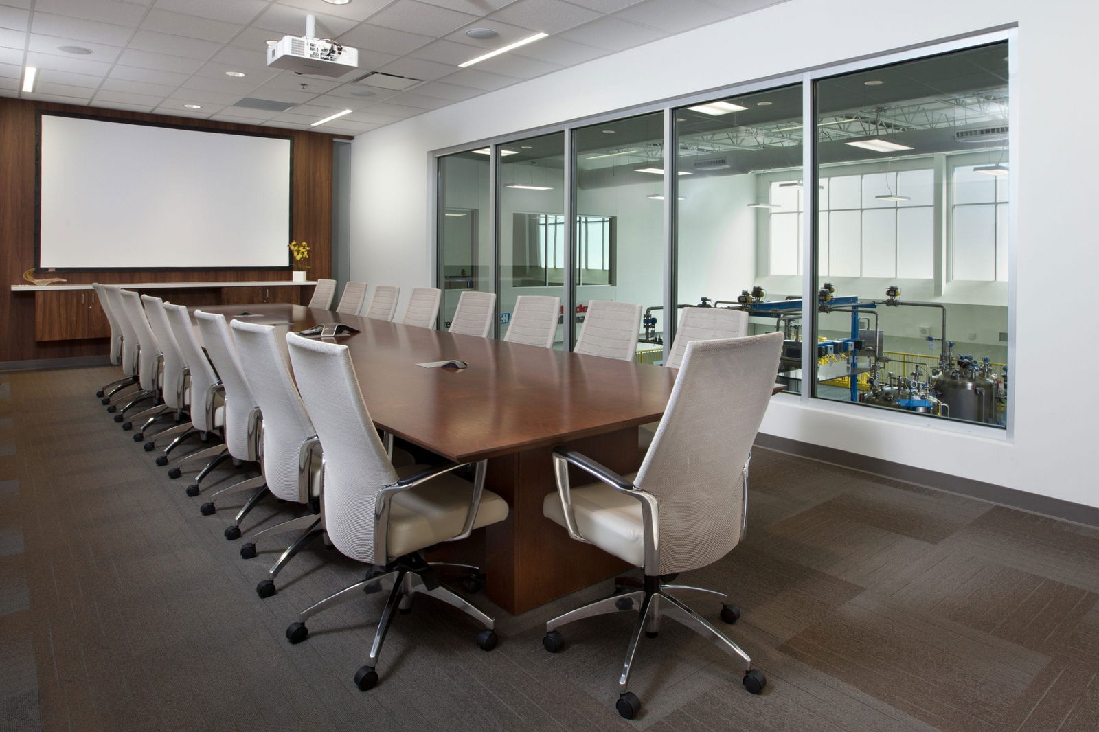 Conference room in the Endress+Hauser Customer Center featuring a polished wooden table surrounded by ergonomic white office chairs, a ceiling-mounted projector and presentation screen with wood-paneled accent wall, and large glass windows overlooking a technical facility with blue equipment and instrumentation, creating an ideal space for business meetings that combines professional aesthetics with functional visibility of the company's operational areas.