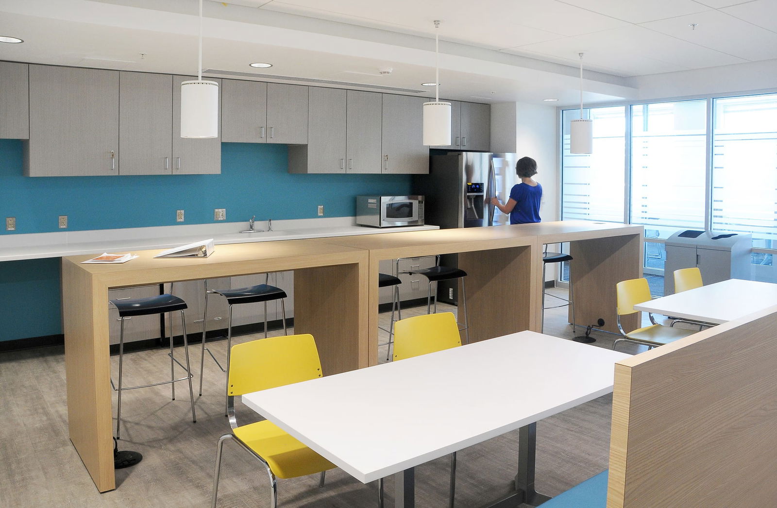 The image shows a modern office kitchen and break area featuring light gray upper cabinets with a bright turquoise blue backsplash. The space includes a kitchen counter with appliances including a microwave and refrigerator, pendant lighting, and a wooden island/bar area with black barstools. Several white tables with bright yellow chairs are arranged in the dining section. A person in a blue shirt is standing by the refrigerator, and the space has large windows providing natural light, creating a contemporary and collaborative workspace environment.