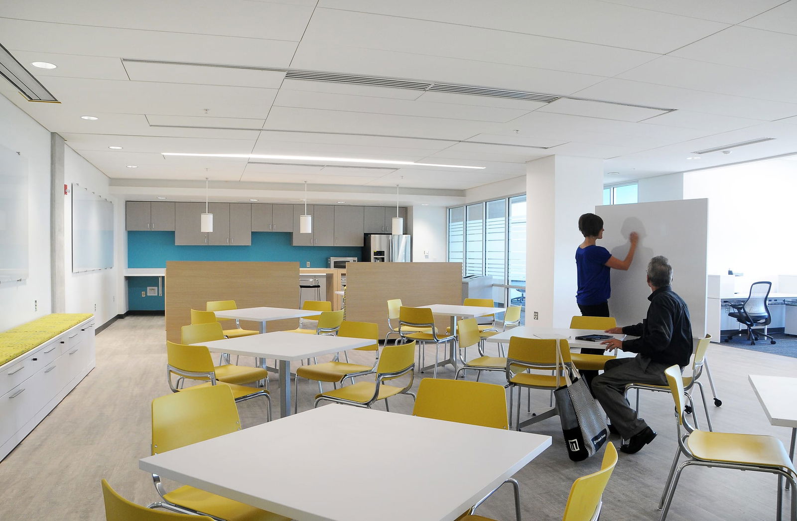 This image shows a bright, modern office break room or collaborative workspace. The space includes several square white tables with bright yellow chairs arranged throughout. The kitchen area in the background has light gray upper cabinets with a distinctive turquoise blue backsplash. A wooden island/counter separates the dining area from the kitchen. On the right side, a person in a blue shirt is writing on a whiteboard while another person in dark clothing is seated at one of the tables with some materials. There's a yellow bench seating along the left wall, large windows providing natural light, and the space has a clean, contemporary design with recessed lighting and pendant fixtures.
