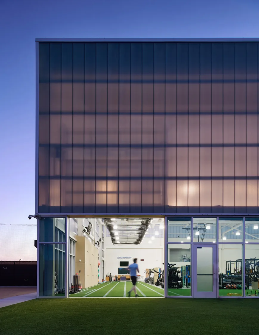 An exterior view looking into an indoor sports training and rehabilitation facility through a garage-style door with artificial green marked with white track lines. The walls consist of floor-to-ceiling windows and garage-style doors, partially open, revealing the interior of the facility. Outside, there is a twilight sky with hues of pink and purple. This photo demonstrates BSA's expertise in architecture, engineering, and designing Sports Medicine facilities.