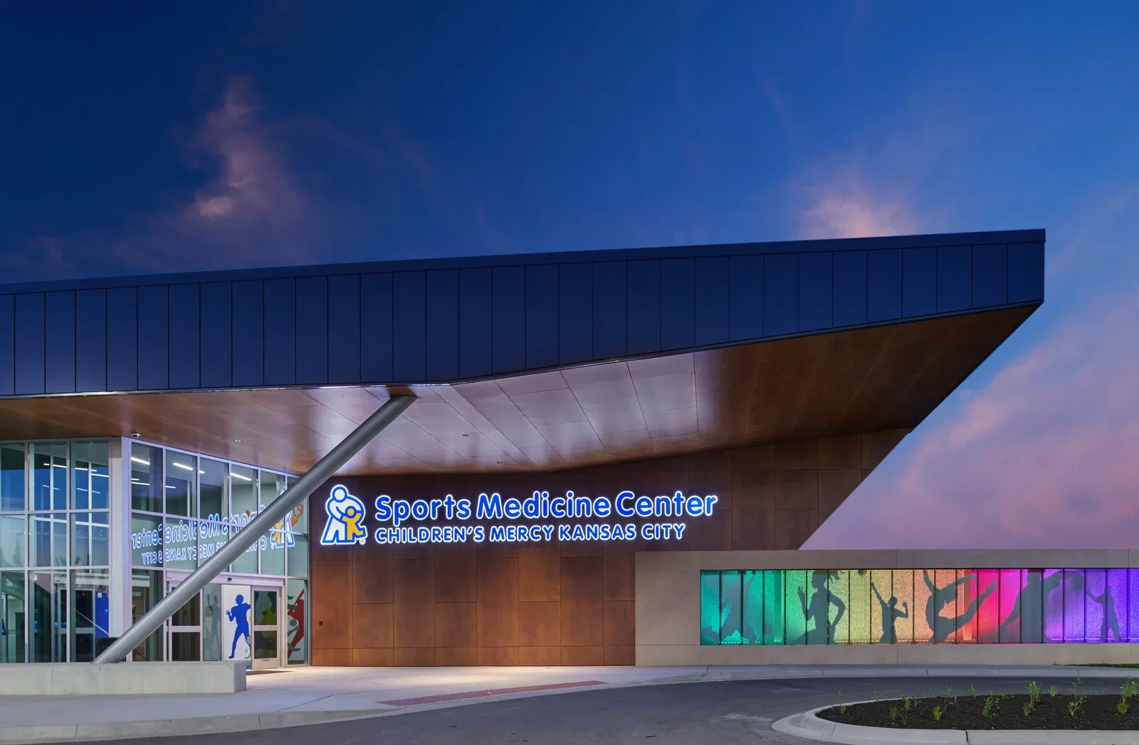 The exterior of the Sports Medicine Center at Children’s Mercy Kansas City during twilight, featuring a modern architectural design with a large angular roof extending outward. The facade is clad in wood paneling, with a prominent illuminated sign displaying the center’s name and logo. A glass entrance with sports-themed graphics is visible on the left. A colorful backlit mural on the right showcases silhouettes of athletes in various dynamic poses, adding a vibrant touch to the facility's sleek and contemporary appearance. This photo demonstrates BSA's expertise in dynamic architectural design.