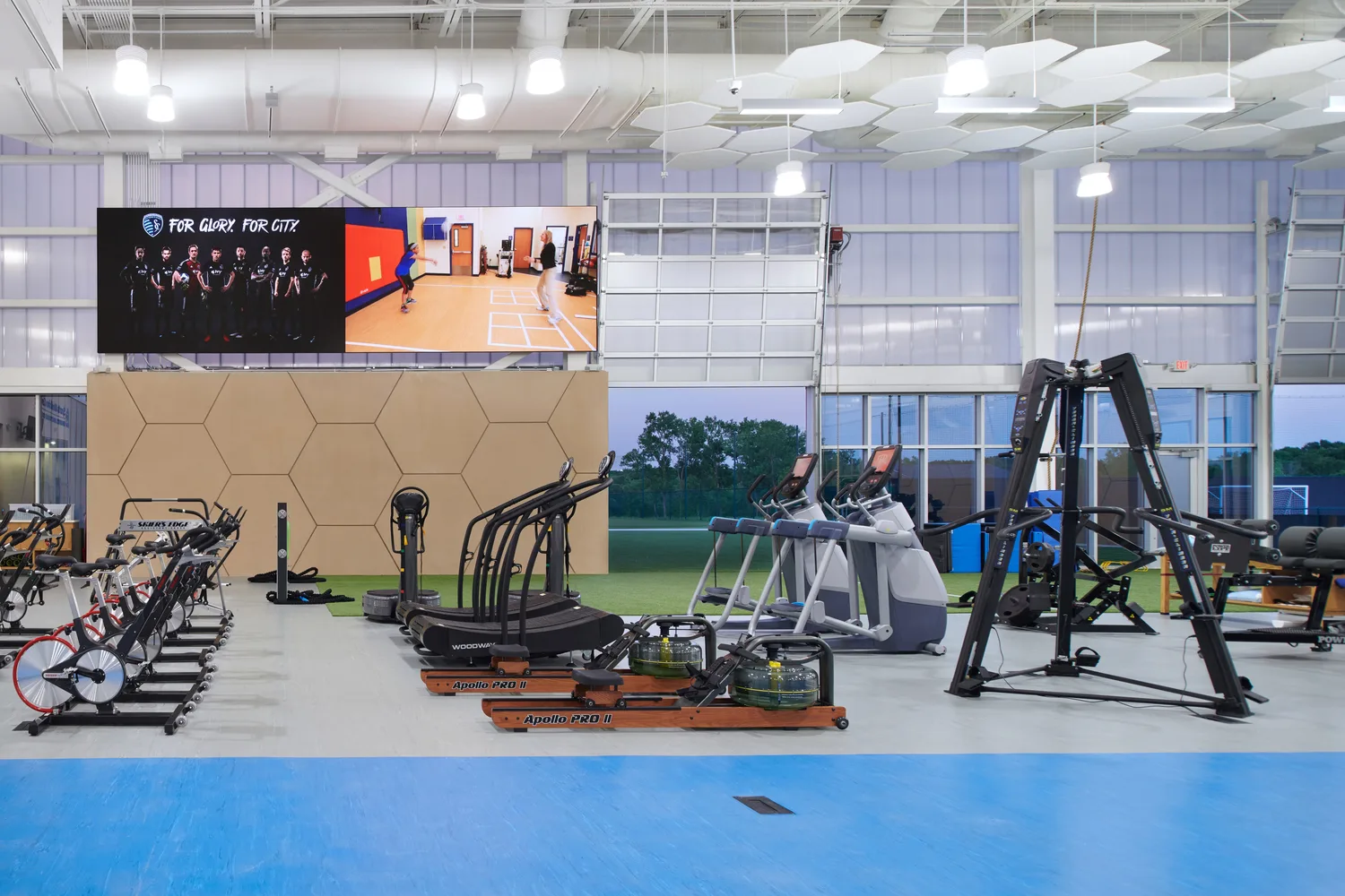 An indoor sports training and rehabilitation facility with various exercise machines arranged in rows. The high-ceiling space features white hexagonal acoustic panels and industrial-style lighting. The far wall consists of floor-to-ceiling windows and garage-style doors, partially open, revealing an outdoor field under a twilight sky with hues of pink and purple. This photo demonstrates BSA's expertise in architecture, engineering, and designing Sports Medicine facilities.
