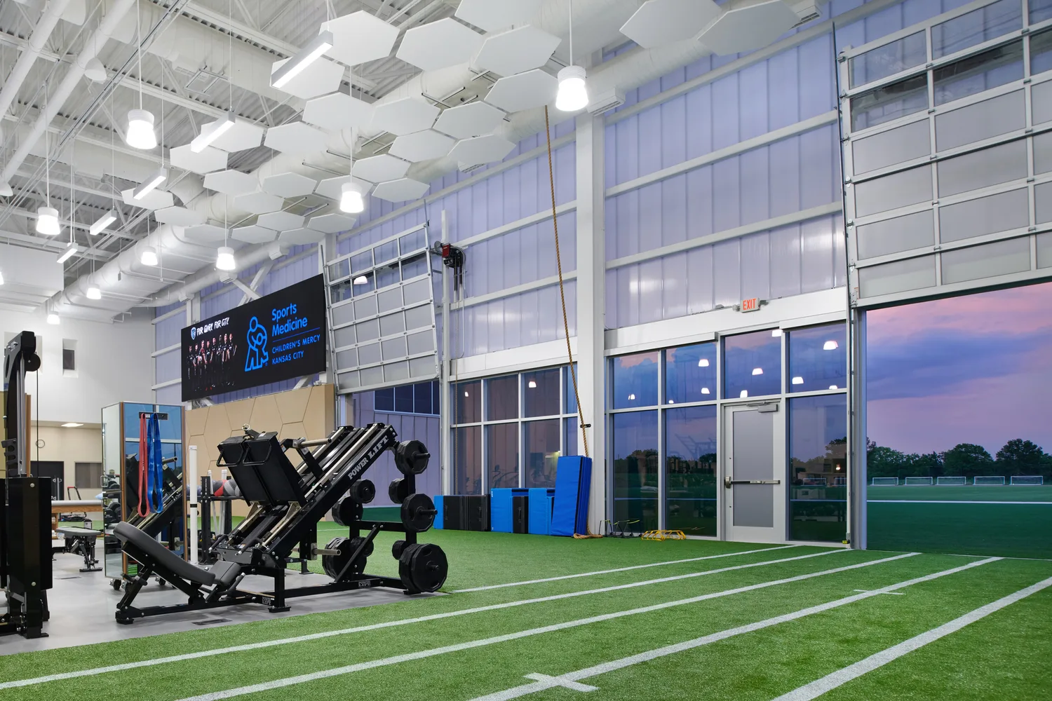 An indoor sports training and rehabilitation facility with artificial turf flooring marked with running lanes. The high-ceiling space features white hexagonal acoustic panels and industrial-style lighting. A leg press machine and resistance training equipment are positioned in the foreground. A large LED screen on the left displays the ‘Sports Medicine - Children’s Mercy Kansas City’ logo. The far wall consists of floor-to-ceiling windows and garage-style doors, partially open, revealing an outdoor field under a twilight sky with hues of pink and purple. This photo demonstrates BSA's expertise in architecture, engineering, and designing Sports Medicine facilities.