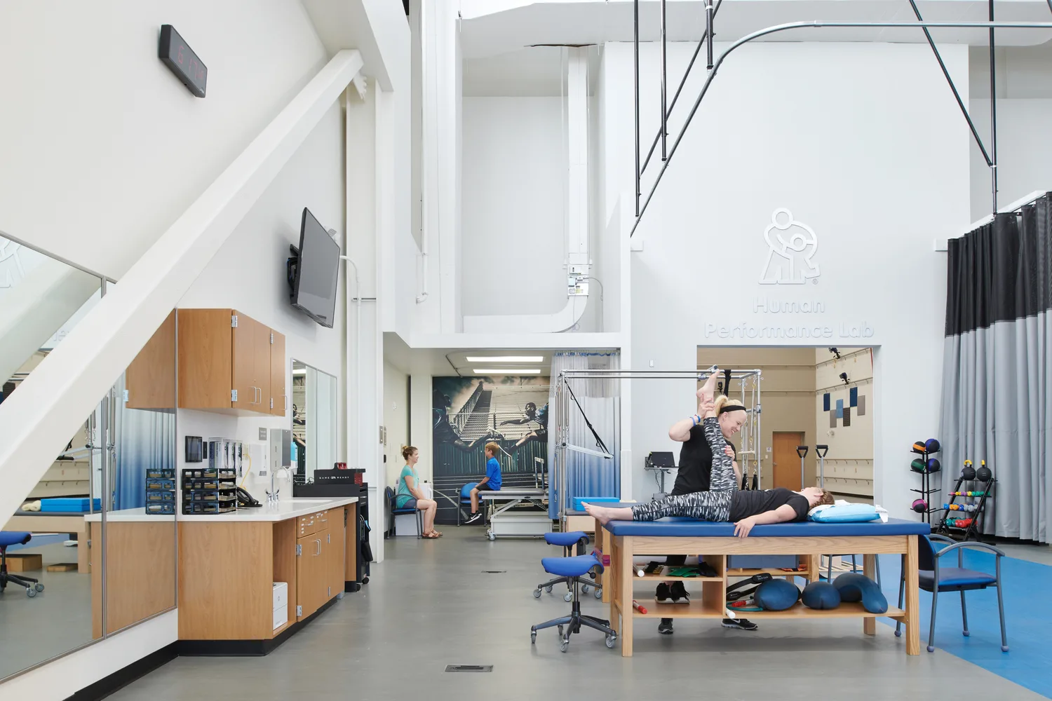 An indoor sports training and rehabilitation facility with a designated area for physical therapy. The high-ceiling space features industrial-style lighting. A woman in exercise clothing is manually stretching the hamstring of a child laying down on blue exam table. Signage above them reads
