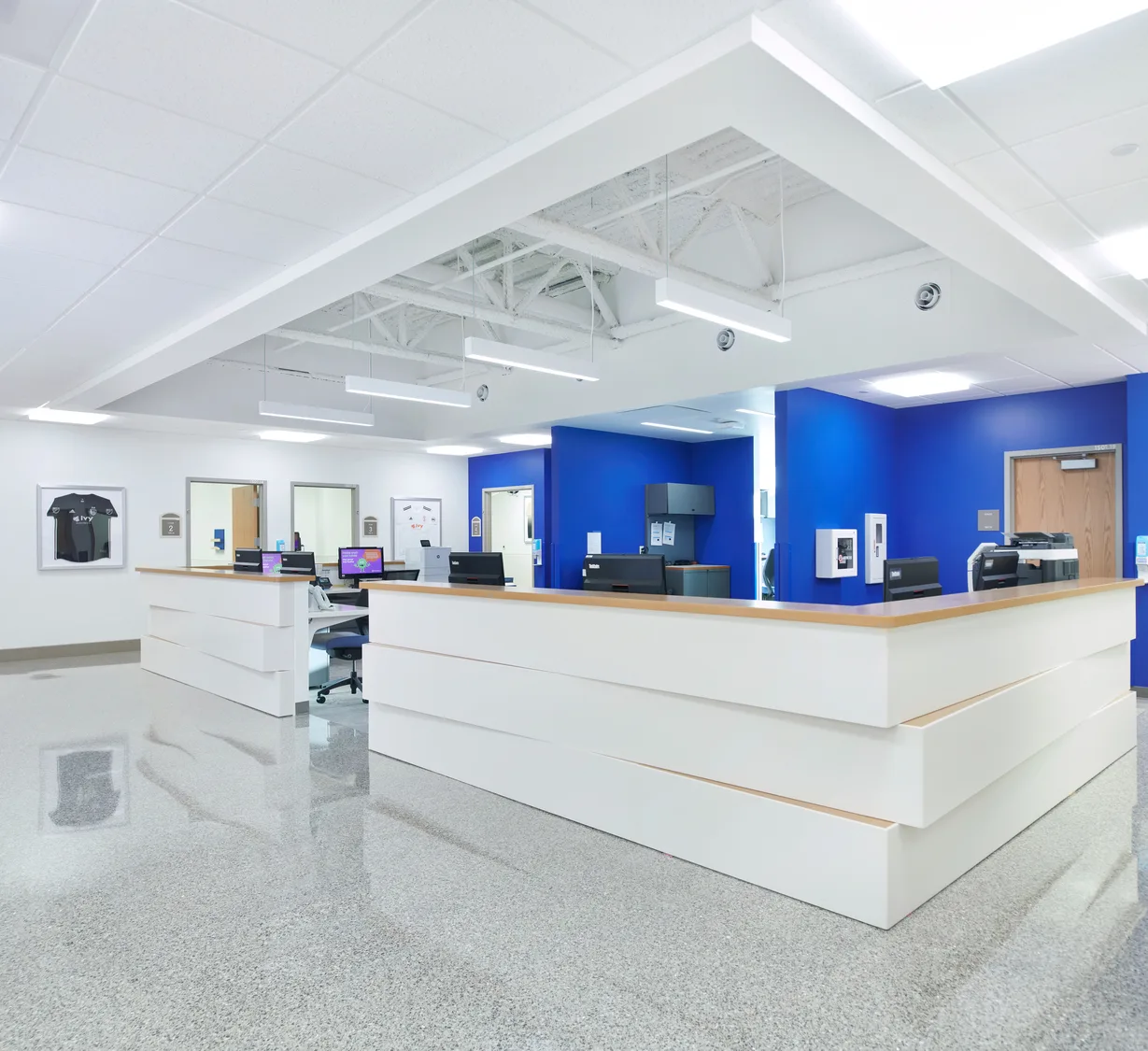 An indoor large U-shaped reception desk in a nearly all-white environment aside from deep royal blue walls behind the desk. The ceiling space features white industrial-style lighting. This photo demonstrates BSA's expertise in interior design as it relates to Sports Medicine facilities.